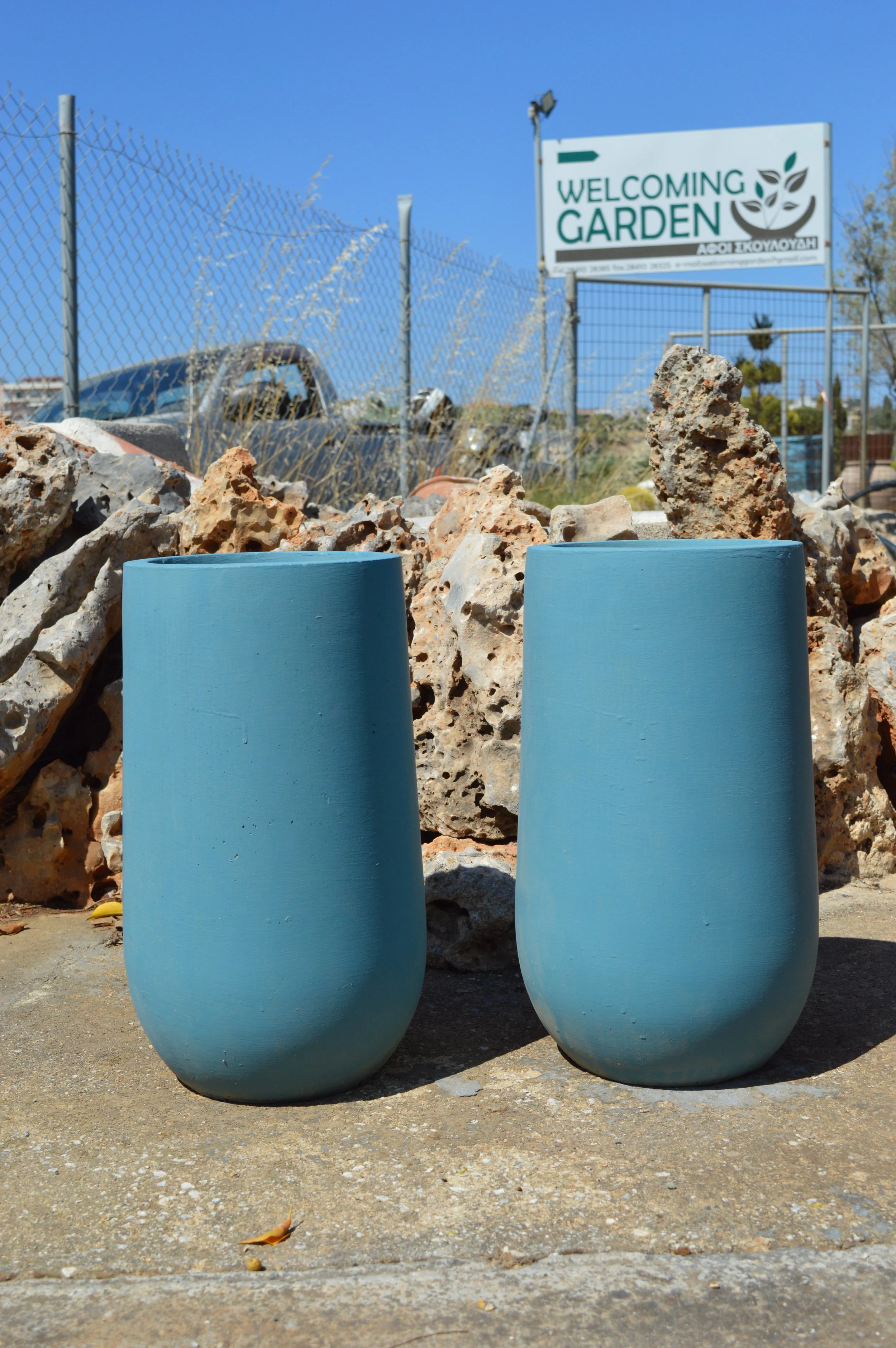 Two large light blue ceramic vases sitting on a sidewalk with a rock and a leaf on the ground. In the background, there is a rock wall, a chain-link fence, a car, and a sign that reads 'Welcoming Garden' with a leaf logo.