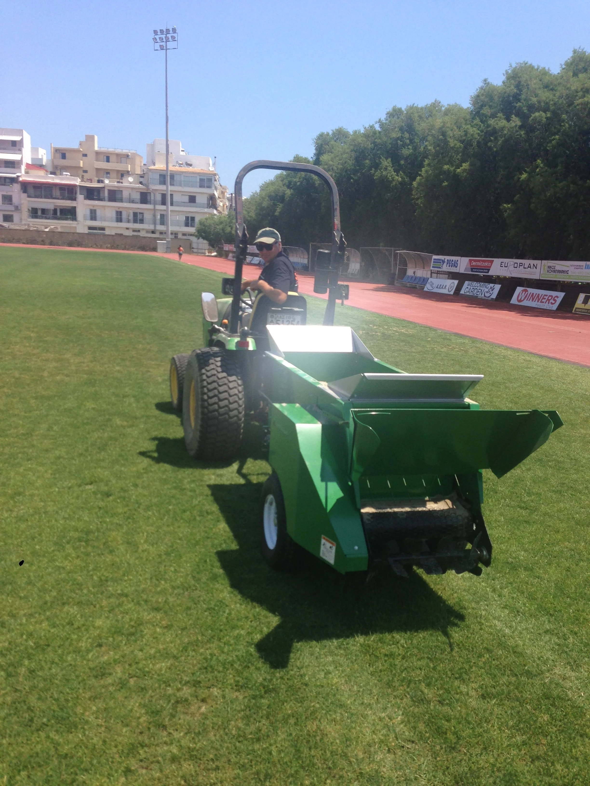 A person operating a green turf maintenance machine on a grass field, with a stadium track and buildings in the background.