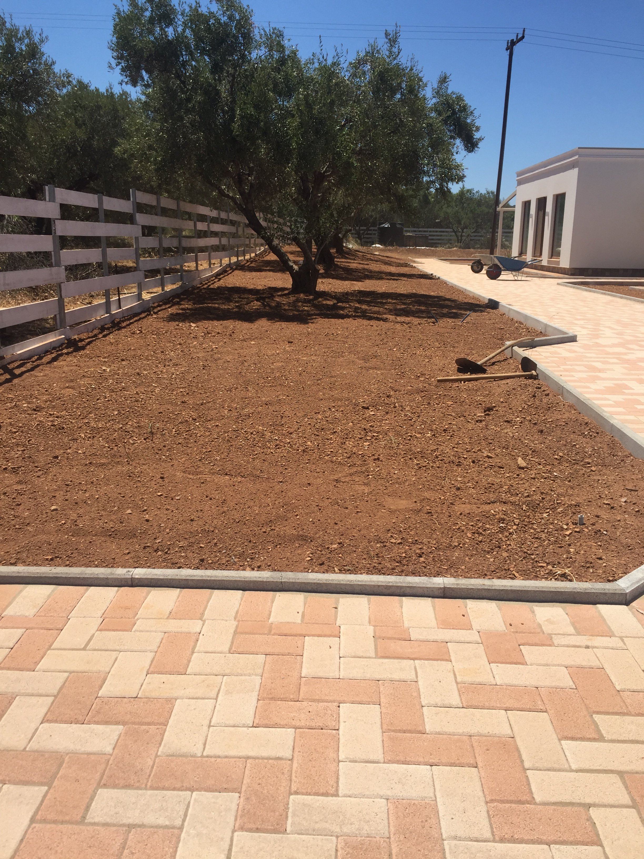 A pathway with newly laid bricks, a wheelbarrow, and construction tools sits adjacent to a white building with large windows. A row of trees and a fence line the background under a clear blue sky.