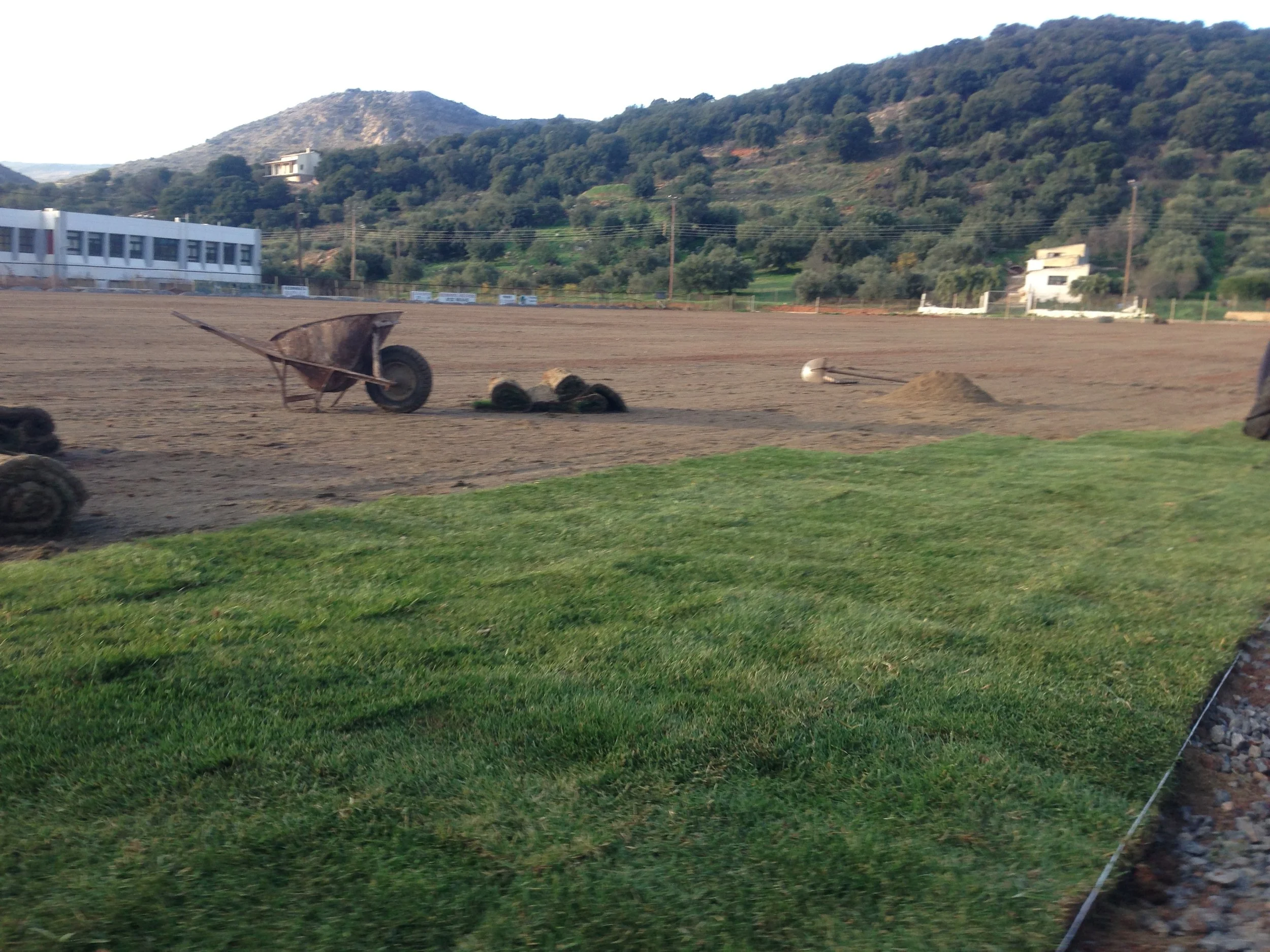 A dirt area with a wheelbarrow, some rocks, and a small pile of dirt; green grass in the foreground and hills with trees and houses in the background.
