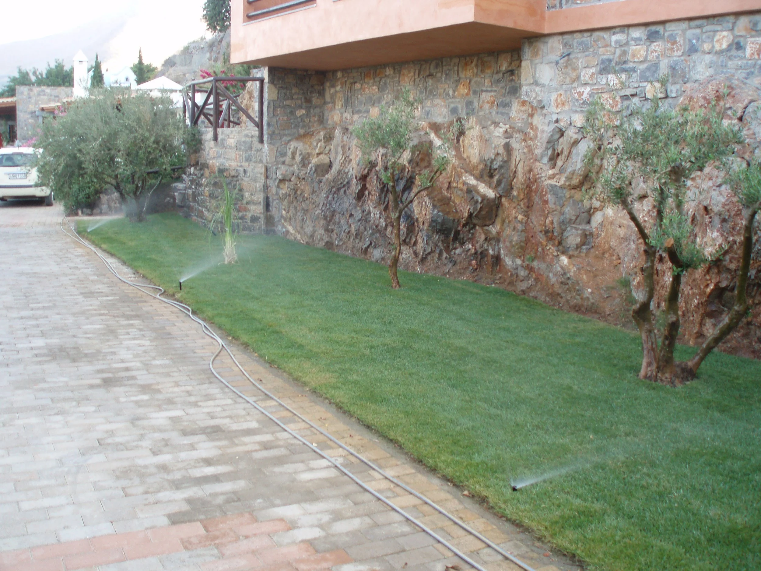 A landscaped yard with green grass being watered by sprinklers, a brick driveway, a stone wall, and trees, with part of a building's balcony visible at the top.