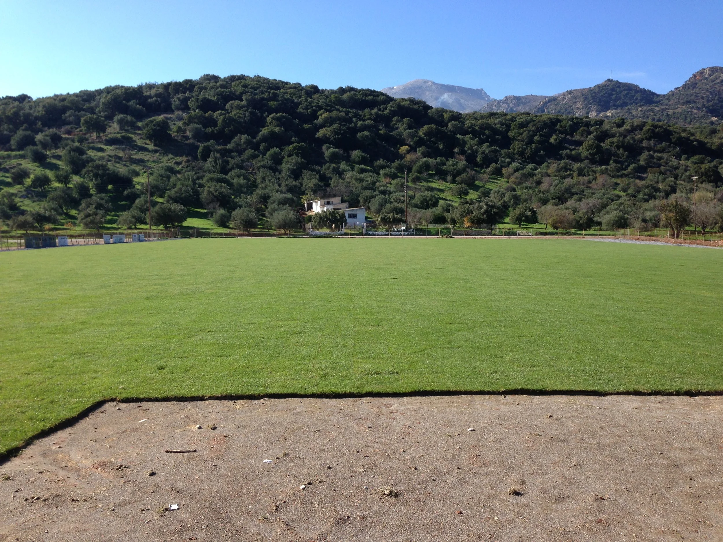 View of a grassy field with a dirt track in the foreground, surrounded by lush green hills dotted with trees, and mountains in the background under a clear blue sky.