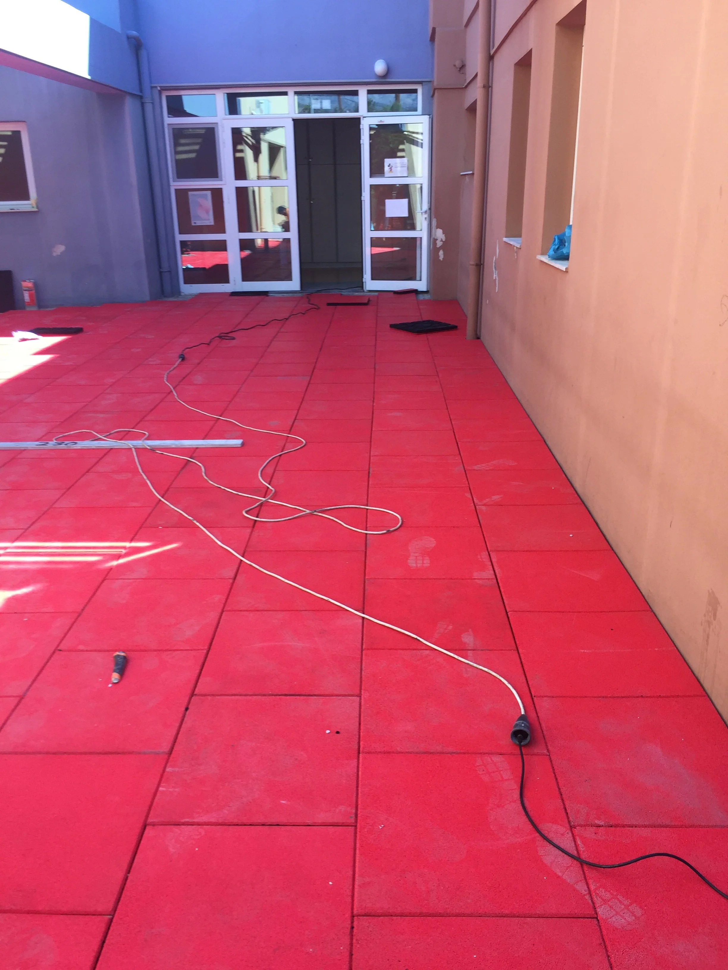 Yellow and gray building exterior with a glass entrance door, red tiles on the ground, and construction wires and tools on the floor.
