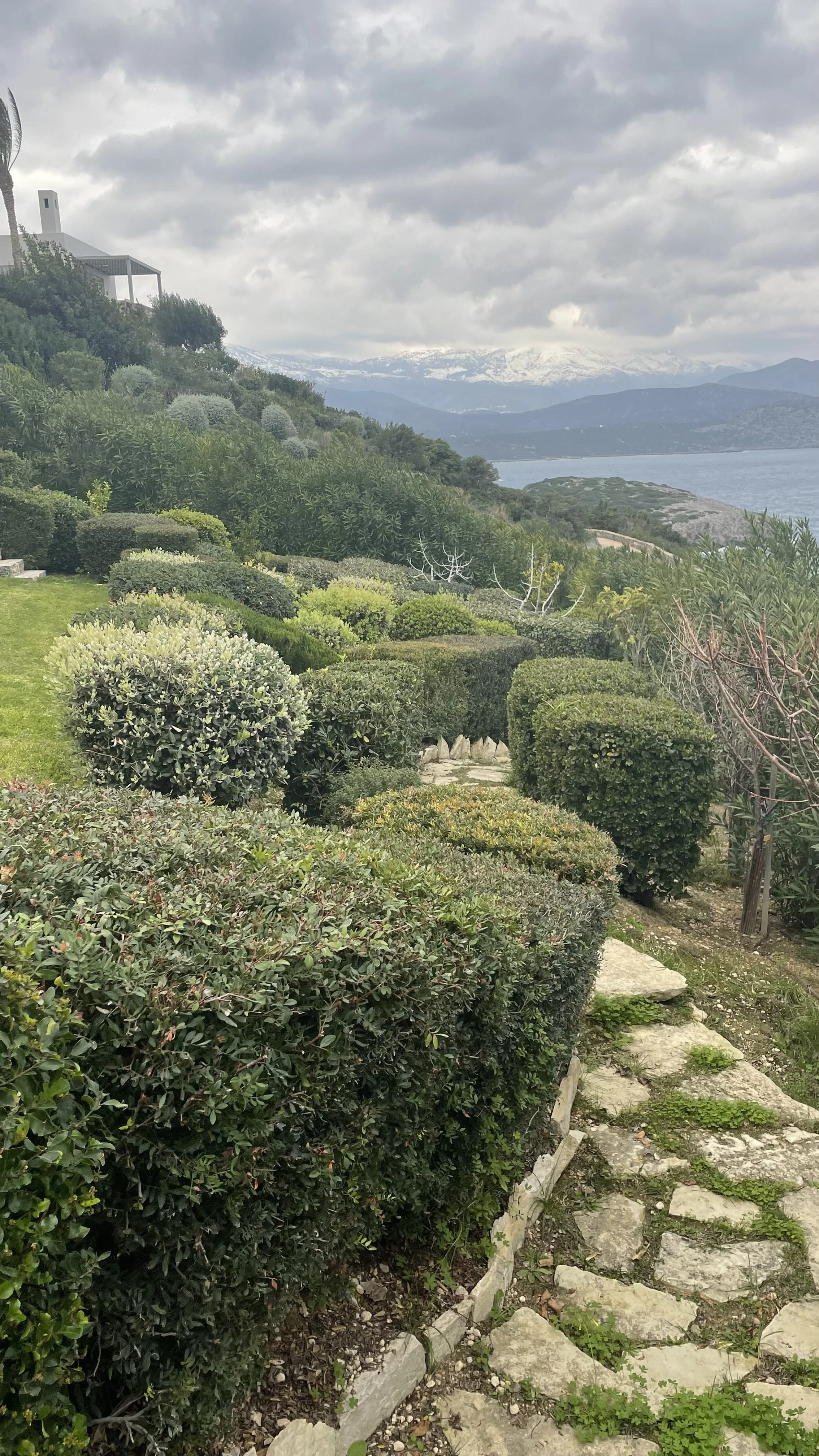 A scenic view of a lush green hillside garden with trimmed bushes, a stone pathway, and a lake with mountains in the distance under cloudy skies.
