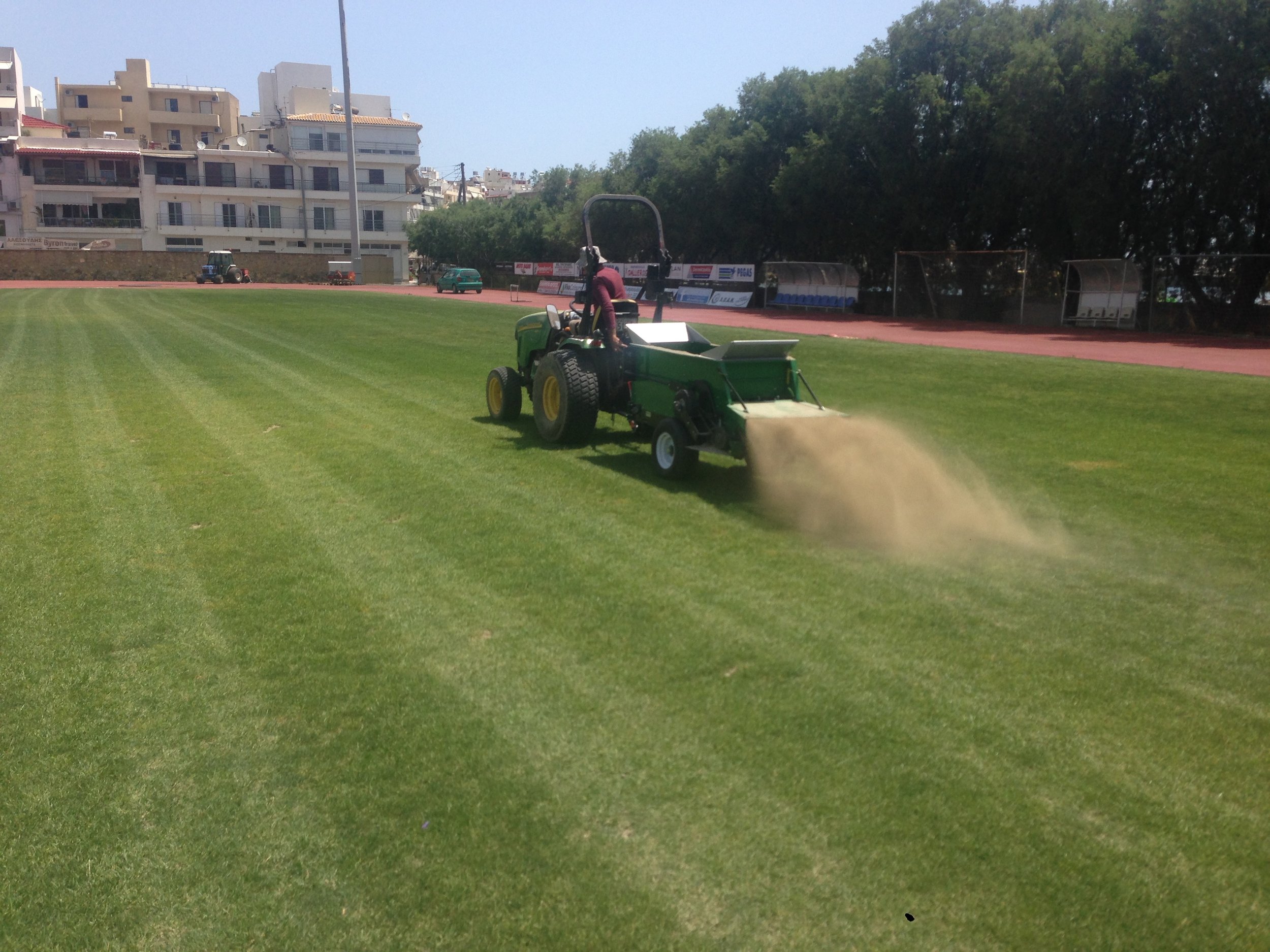 A person operating a green lawn tractor on a sports field, with a grassy area, a running track, and residential buildings with balconies in the background.