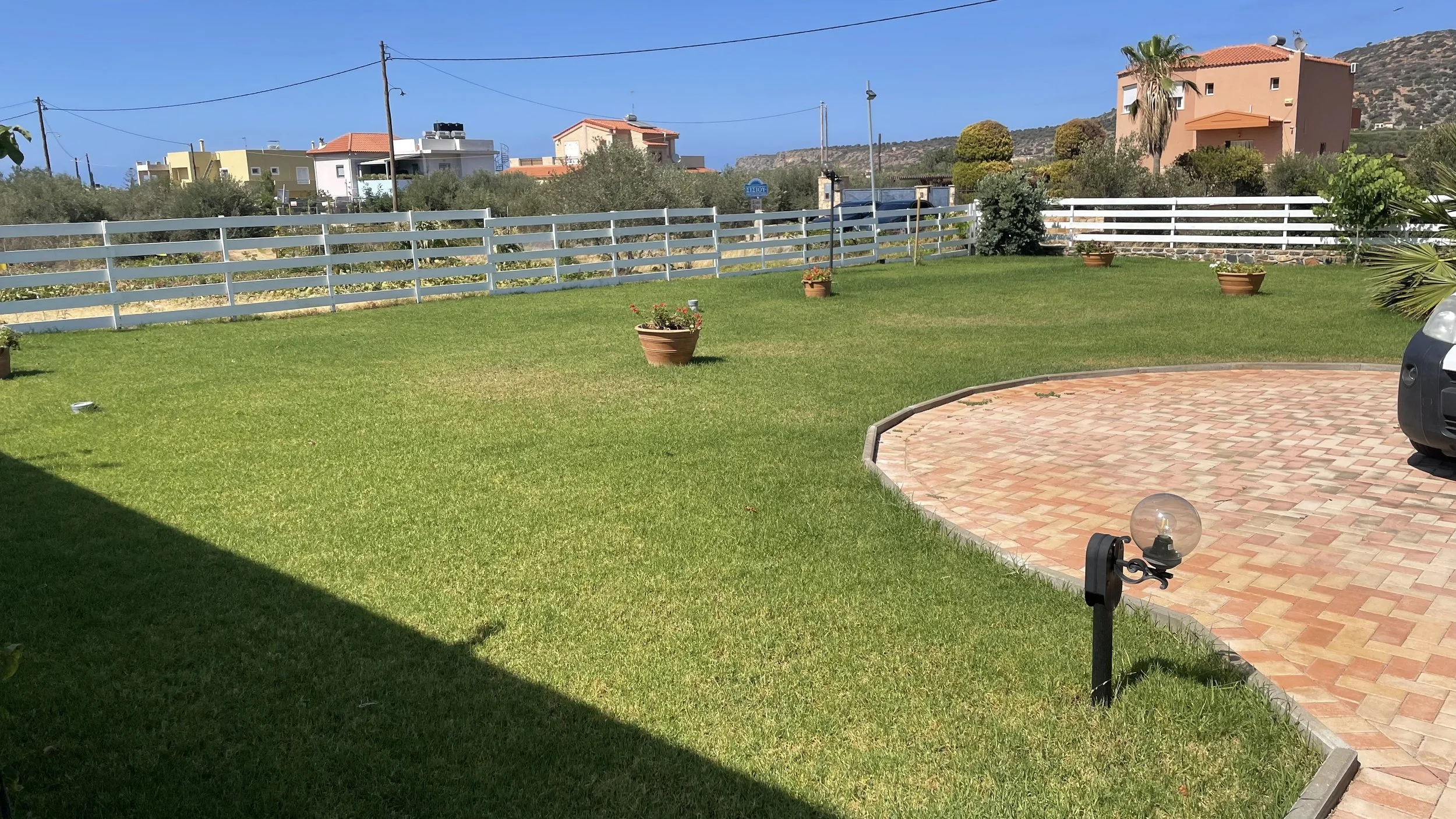 A backyard with a well-maintained lawn, potted plants, a brick patio, and a white fence. Houses and mountainous terrain are visible in the background under a clear blue sky.