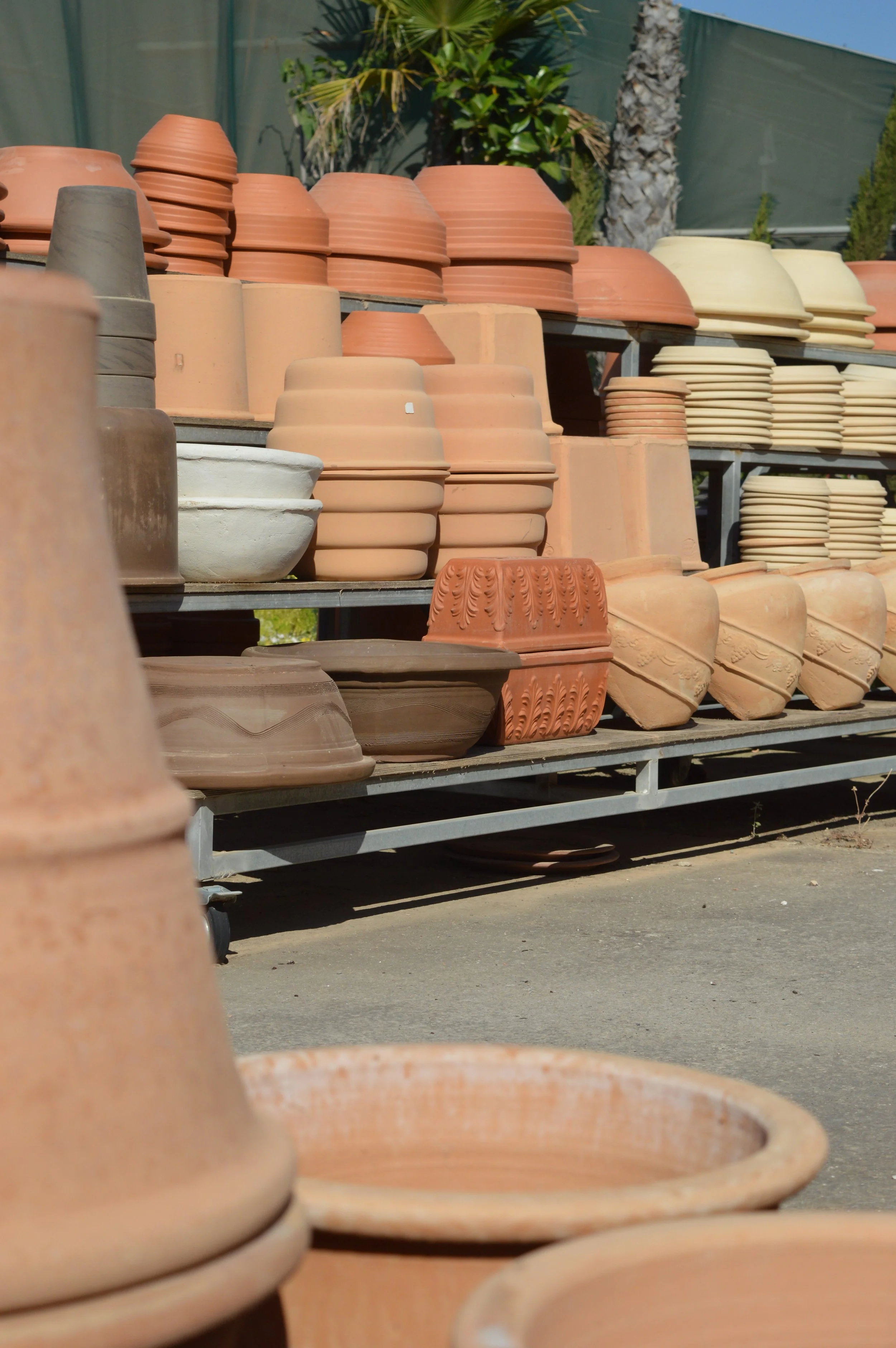 Stacks of terracotta and clay plant pots displayed outdoors on metal shelves, with trees and a fence in the background.