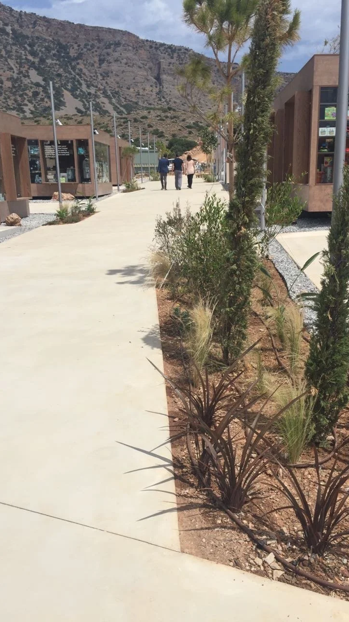 A concrete walkway in a modern outdoor setting with landscaped plants and trees, and a mountainous backdrop, with three people walking in the distance.