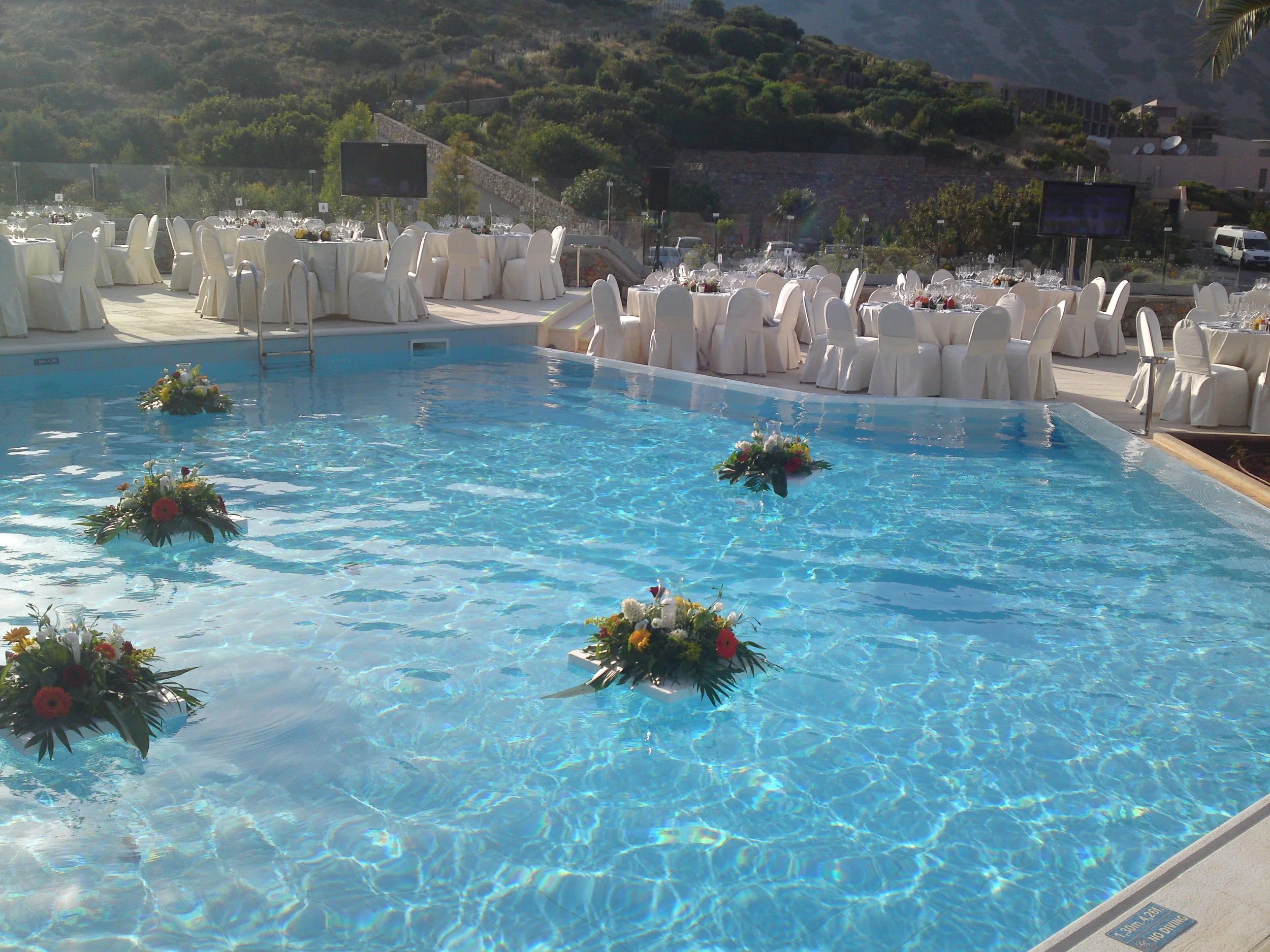 Outdoor event setup near a swimming pool with round tables covered in white tablecloths and white chairs with white covers. The tables are decorated with floral centerpieces. There are two large screens on stands behind the tables. The location is su