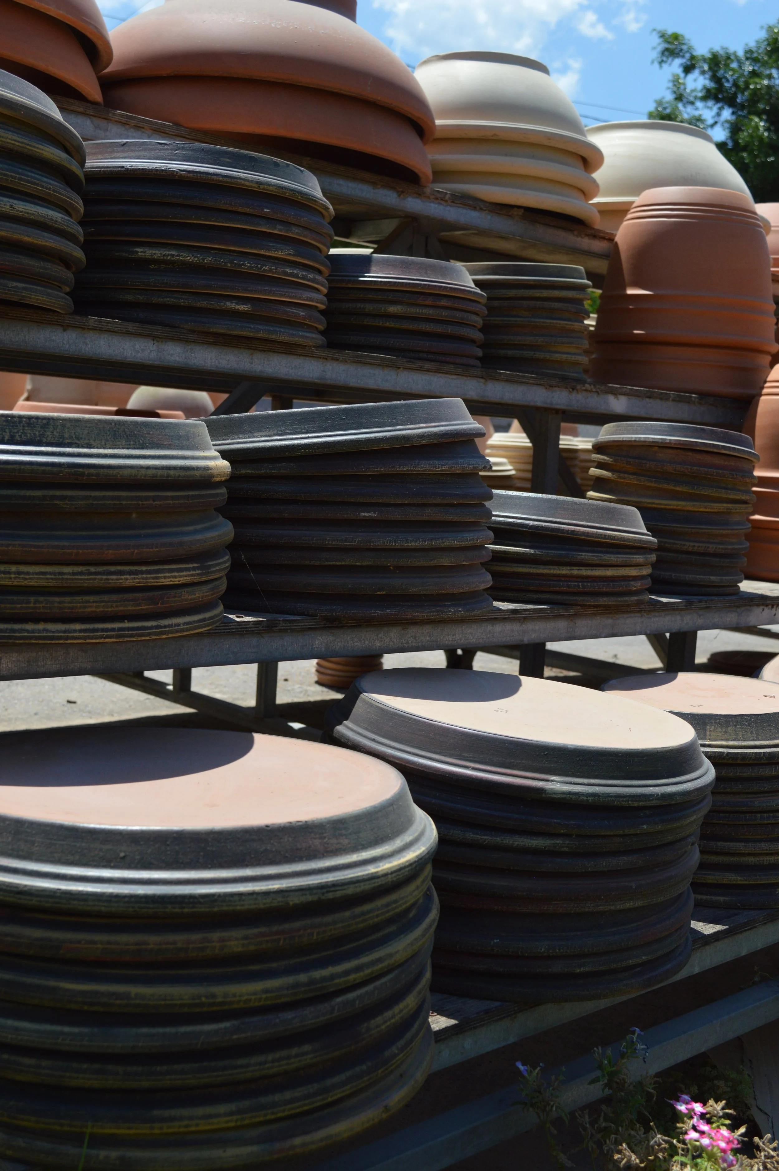Stacks of empty clay flowerpots on metal racks outdoors with blue sky and green trees in the background.