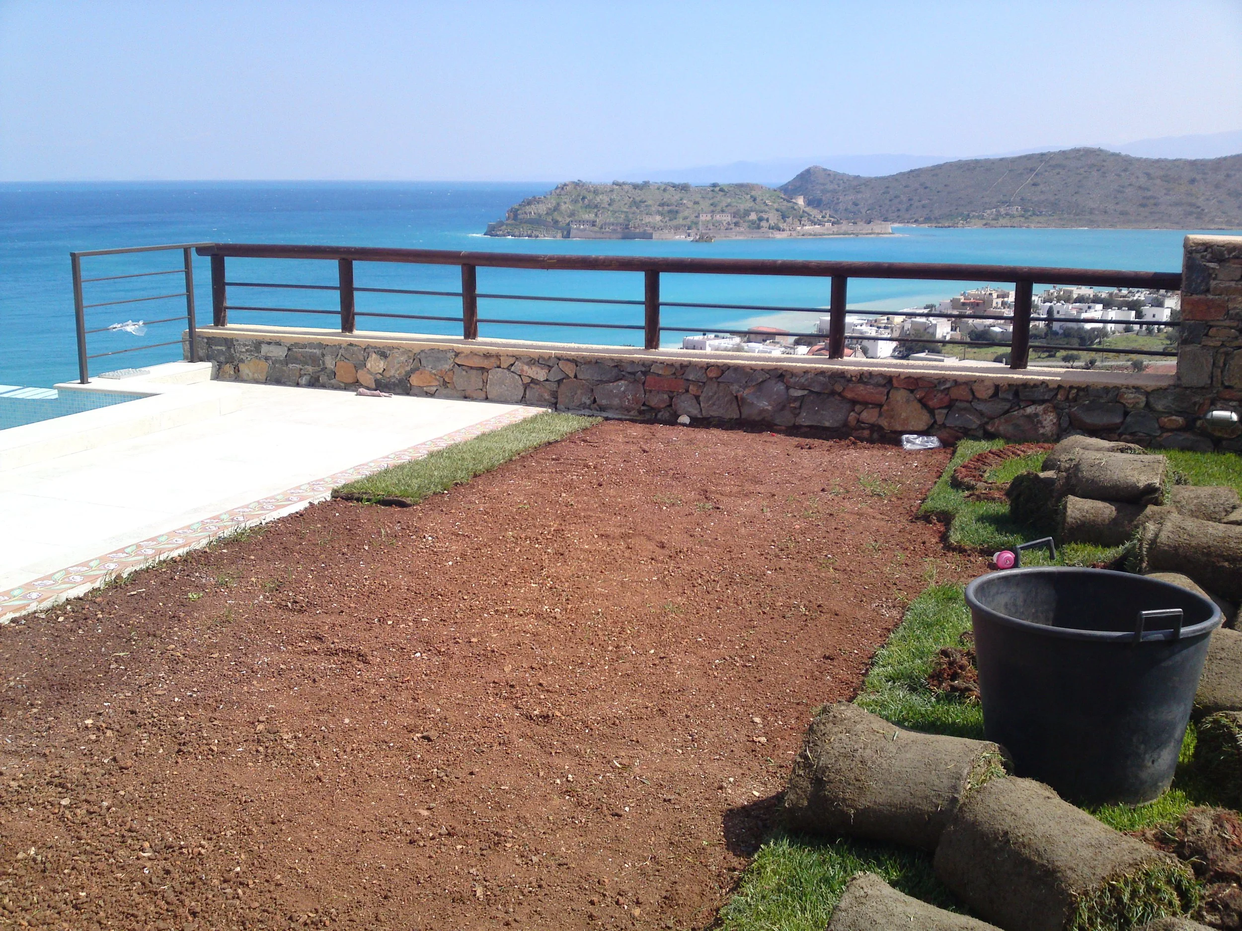 A view of a coastal landscape with a stone and wood fence, a dirt patch, a black bucket, and a view of blue ocean and hills in the distance.