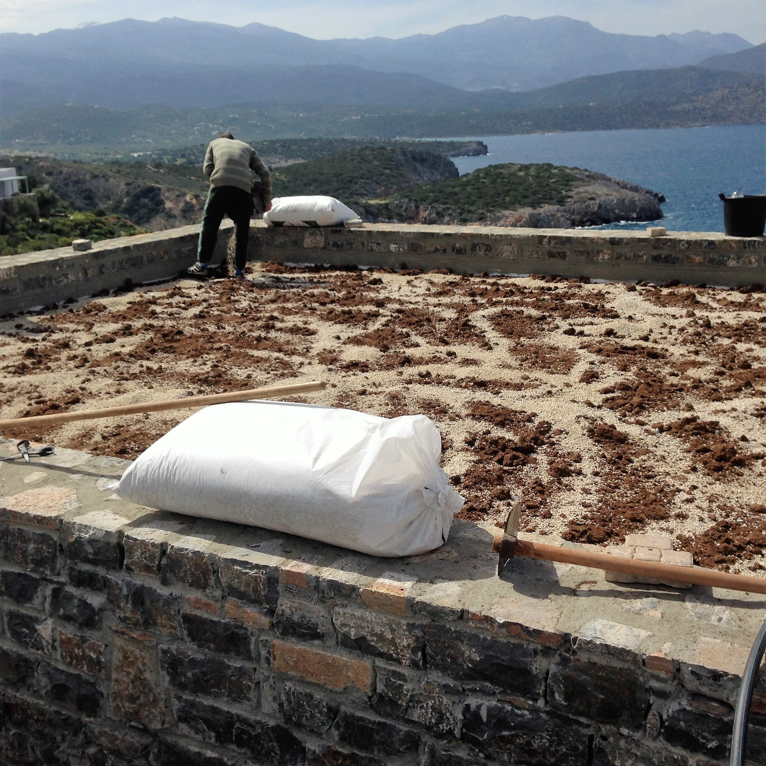 A person wearing a jacket and black pants working on a construction project outdoors with a view of mountains, a body of water, and hilly terrain in the background.