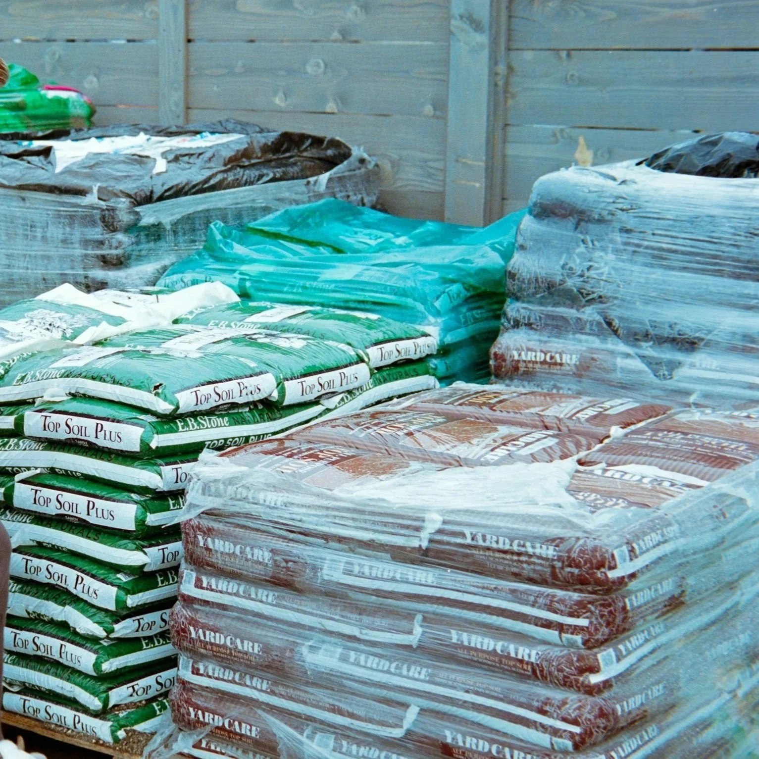 Stacks of bags containing gardening soil and yard care products, wrapped in plastic, arranged outdoors against a wooden fence.