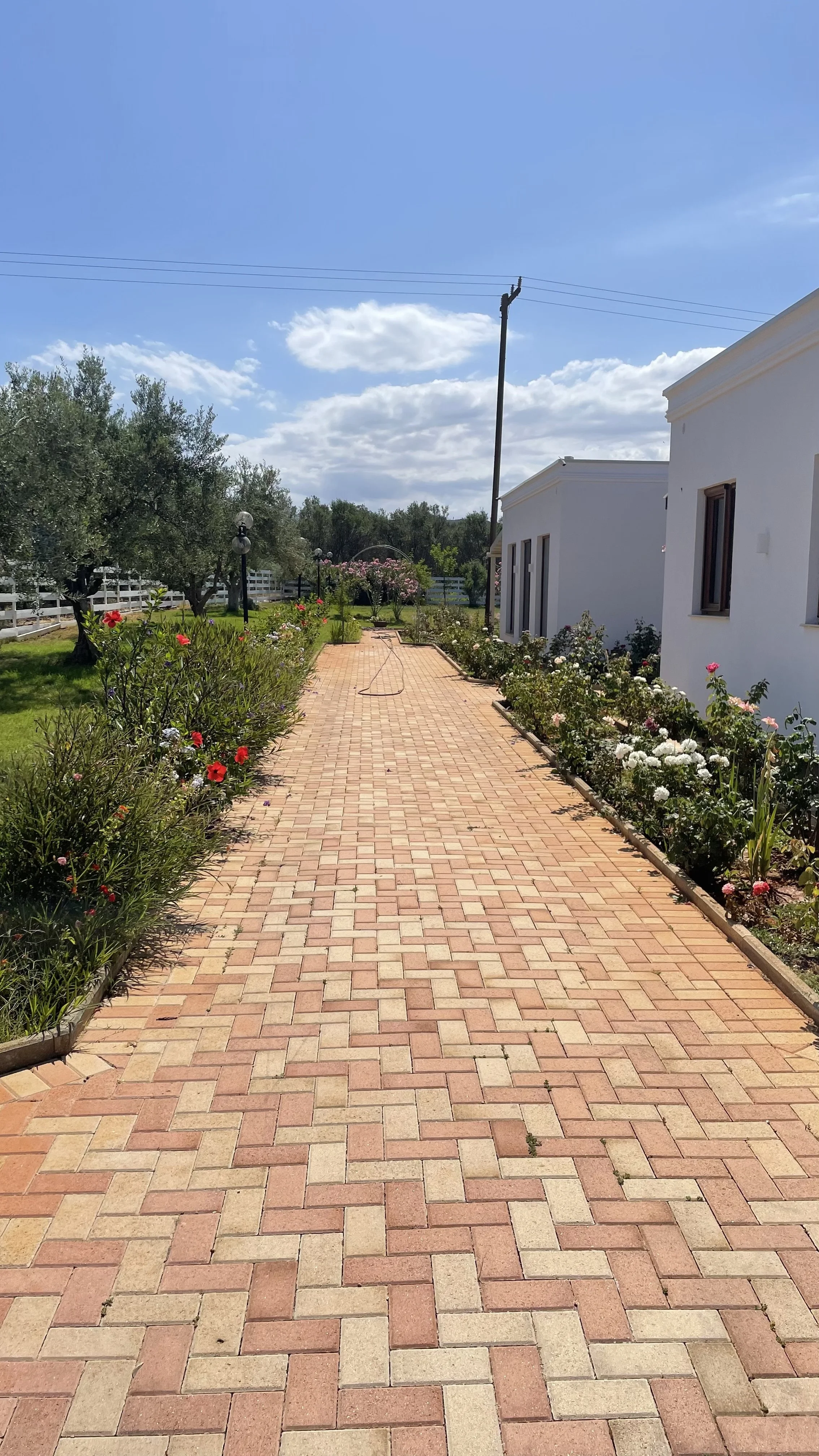 A brick pathway lined with colorful flowers and bushes, leading to a garden area with trees and a white fence, under a partly cloudy blue sky.