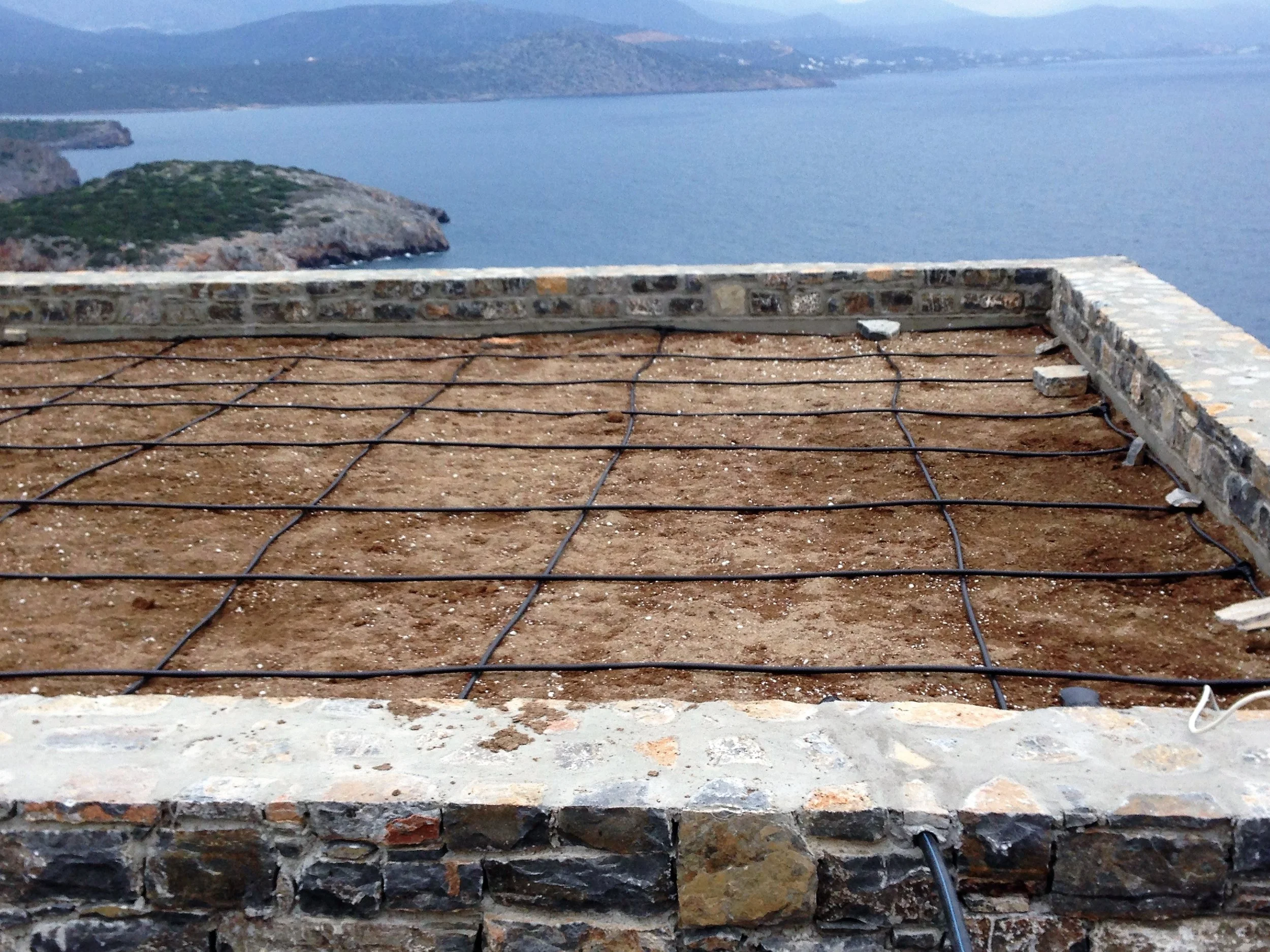View of a rocky coastline with a body of water in the background, taken from a stone-walled balcony under construction with exposed rebar and construction materials.