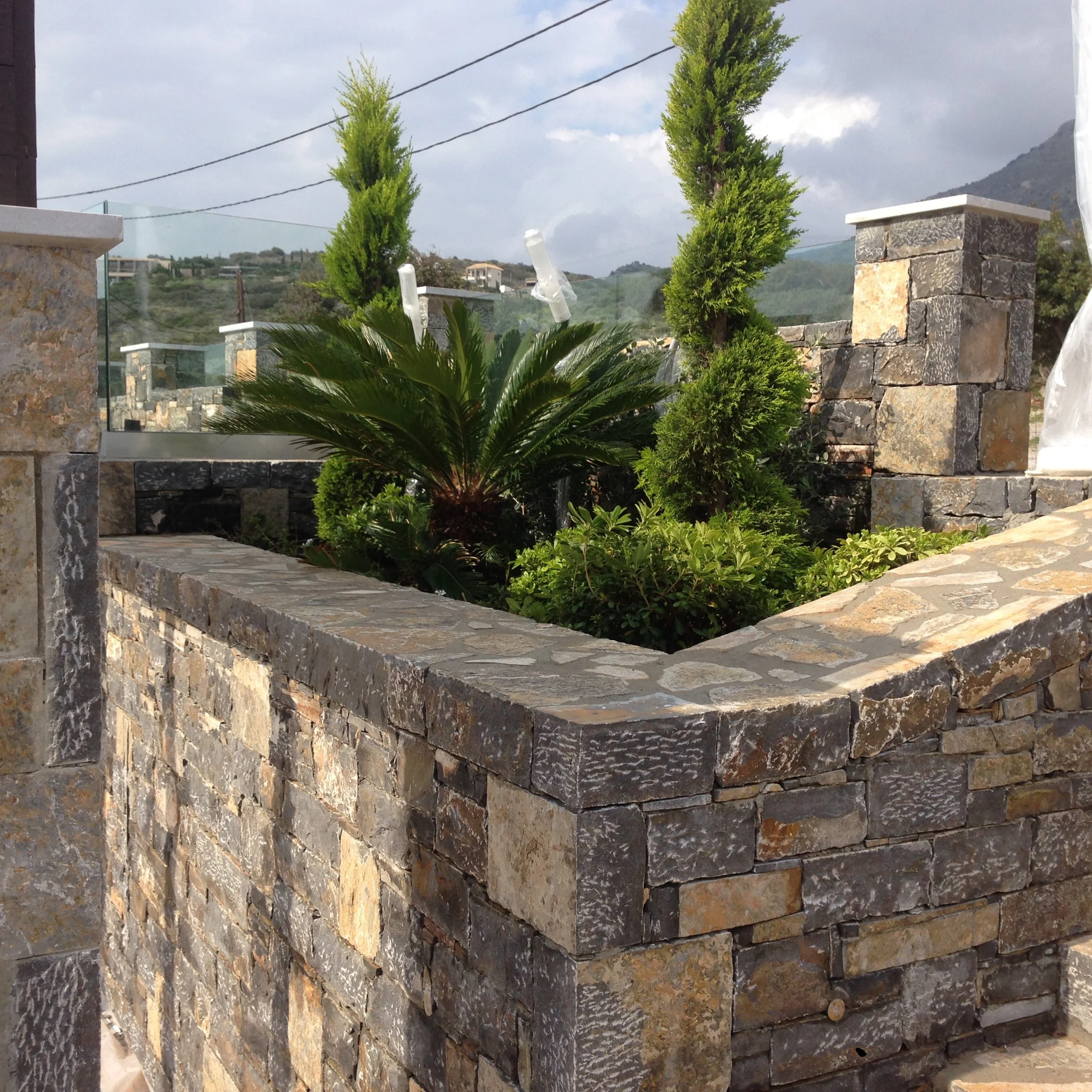 A view of an outdoor stone courtyard with bushes, two tall green trees, and a stone wall, with hills and houses in the background and utility wires overhead.
