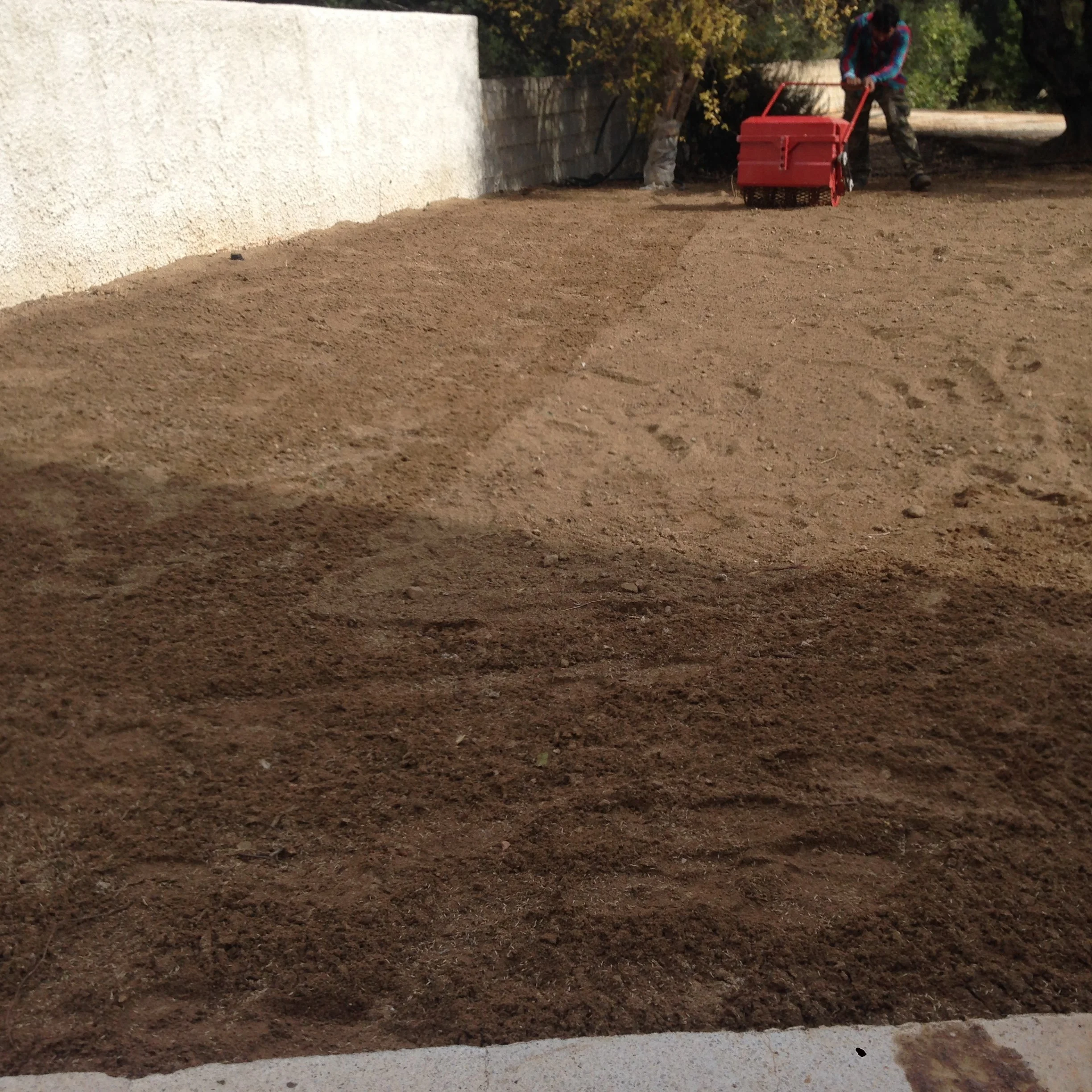 Person dressed in camouflage pants and a blue plaid shirt is pushing a red lawn aerator on a dirt surface next to a concrete wall and some trees.