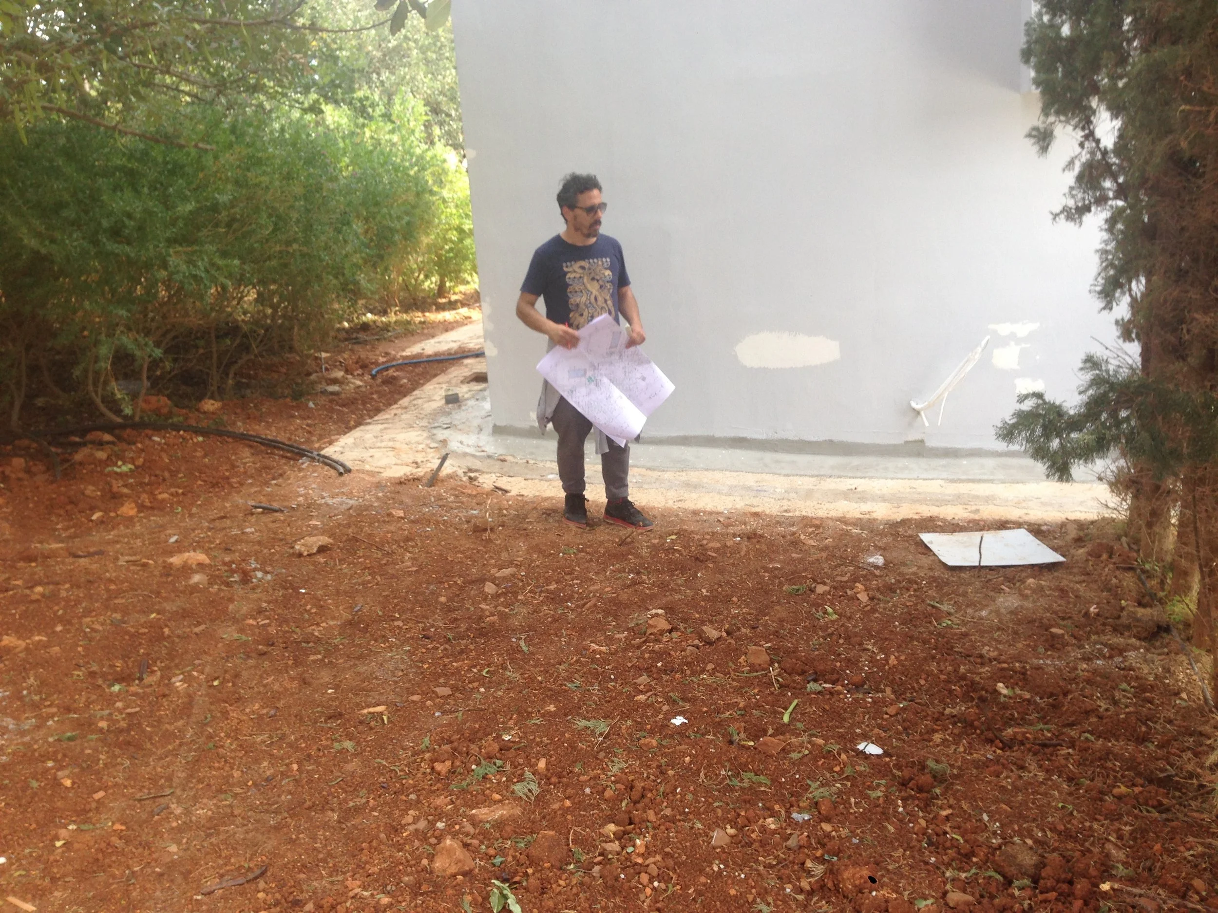 A man standing outdoors on a dirt path, holding architectural plans, near greenery, with a white building or wall behind him.