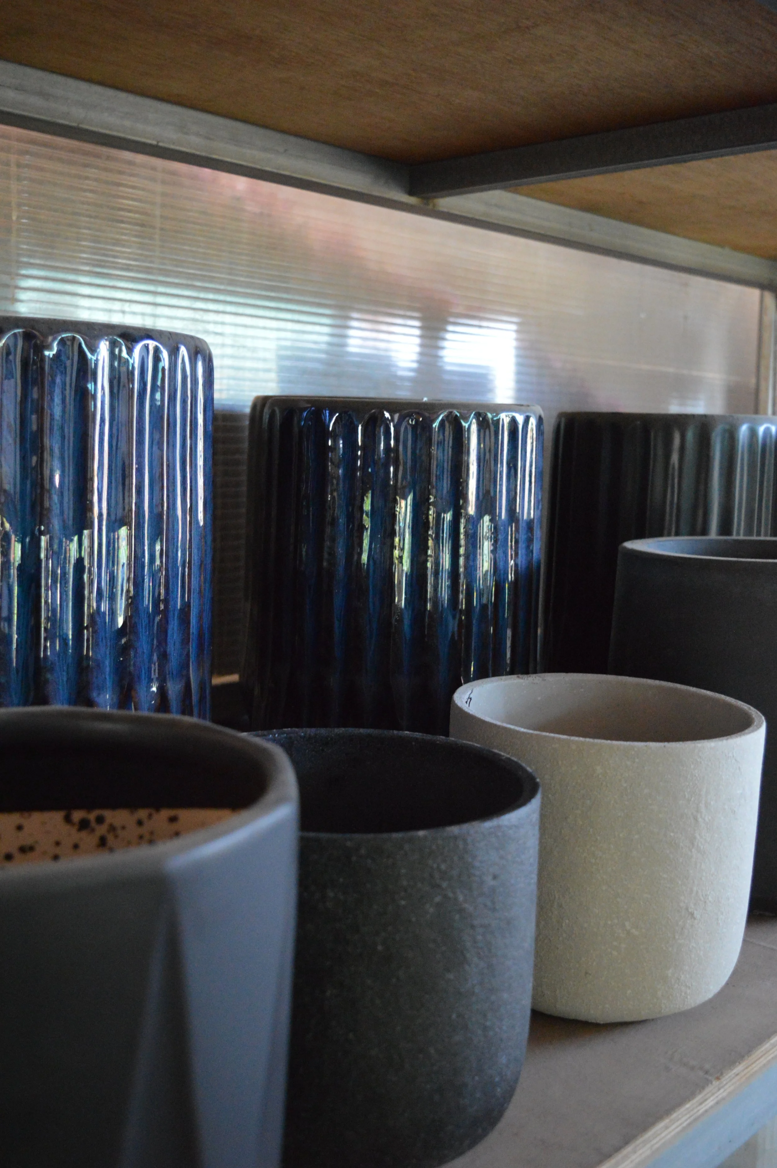 Various ceramic cups with different colors and textures arranged on a shelf with a reflective metallic backsplash and wooden ceiling above.