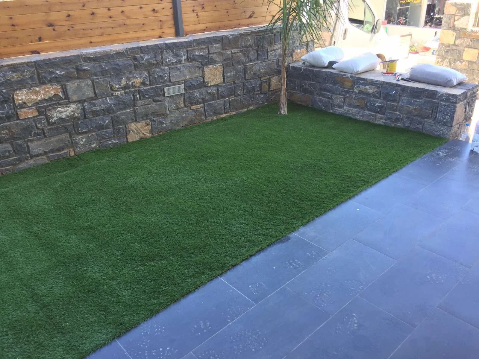 Partial view of a backyard patio featuring artificial grass, a stone wall with a wooden fencing above, a small tree, and some construction supplies on a stone ledge.