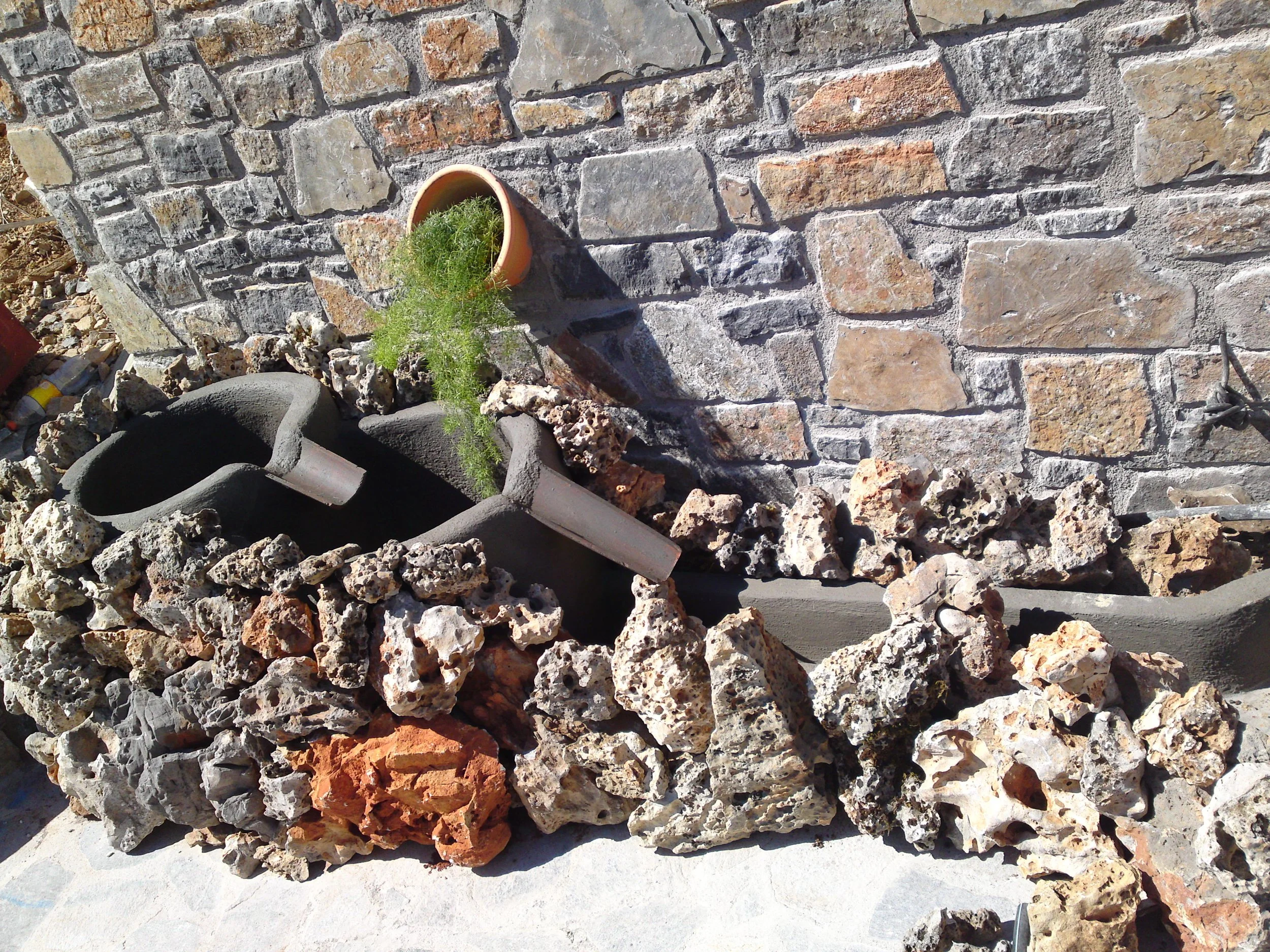 Close-up of a brick and stone wall with a small flower pot and some tools at the bottom.