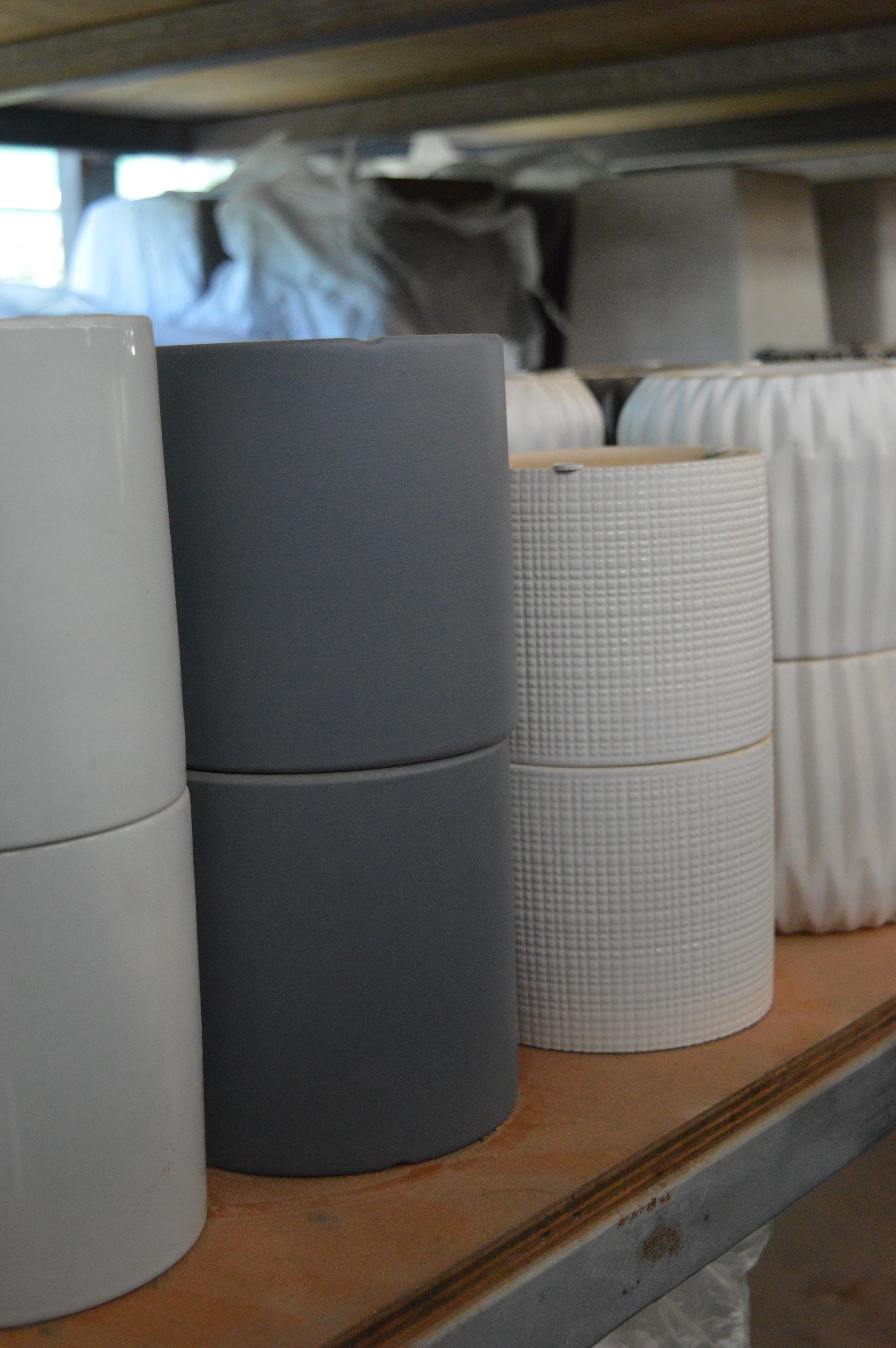 Stacked cylindrical containers on a wooden shelf, some are plain white, others are textured or grey, with a fabric-like material and a box in the background.