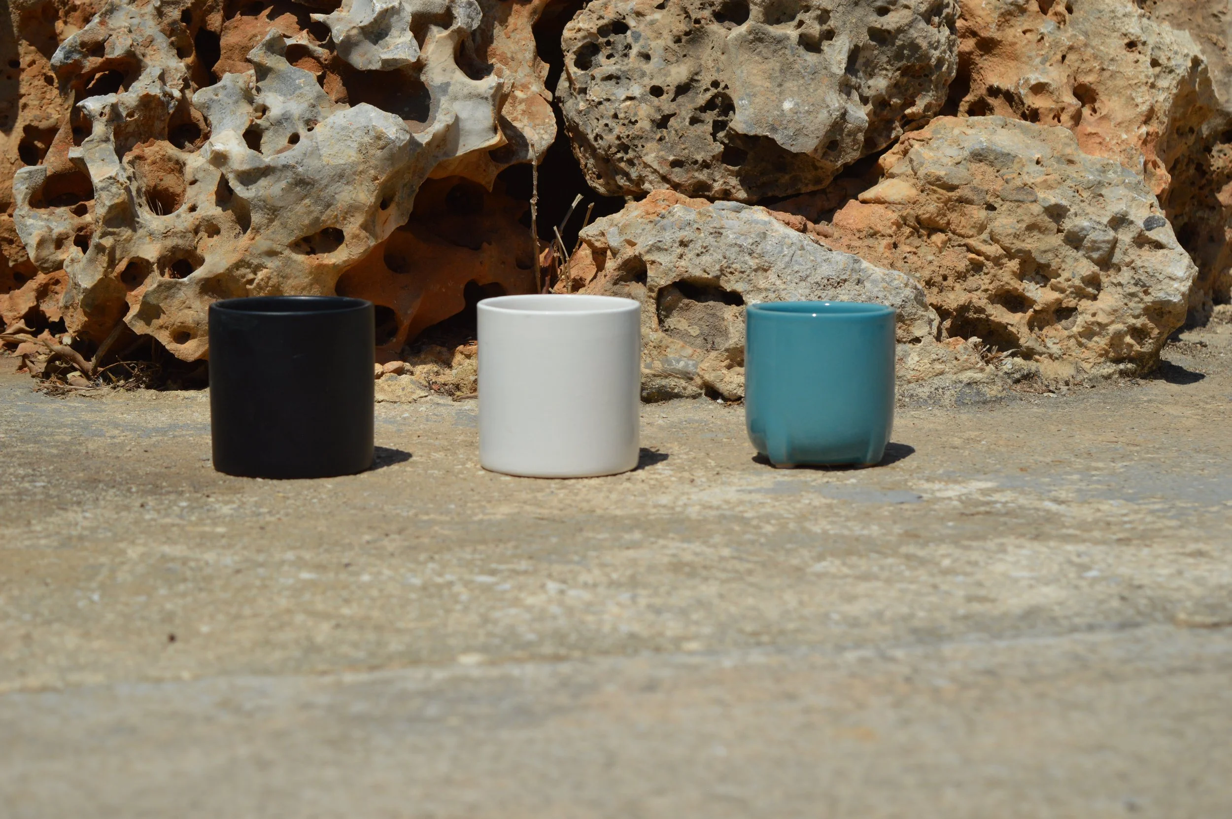 Three ceramic cups in black, white, and turquoise colors placed on sandy ground in front of a rocky background.