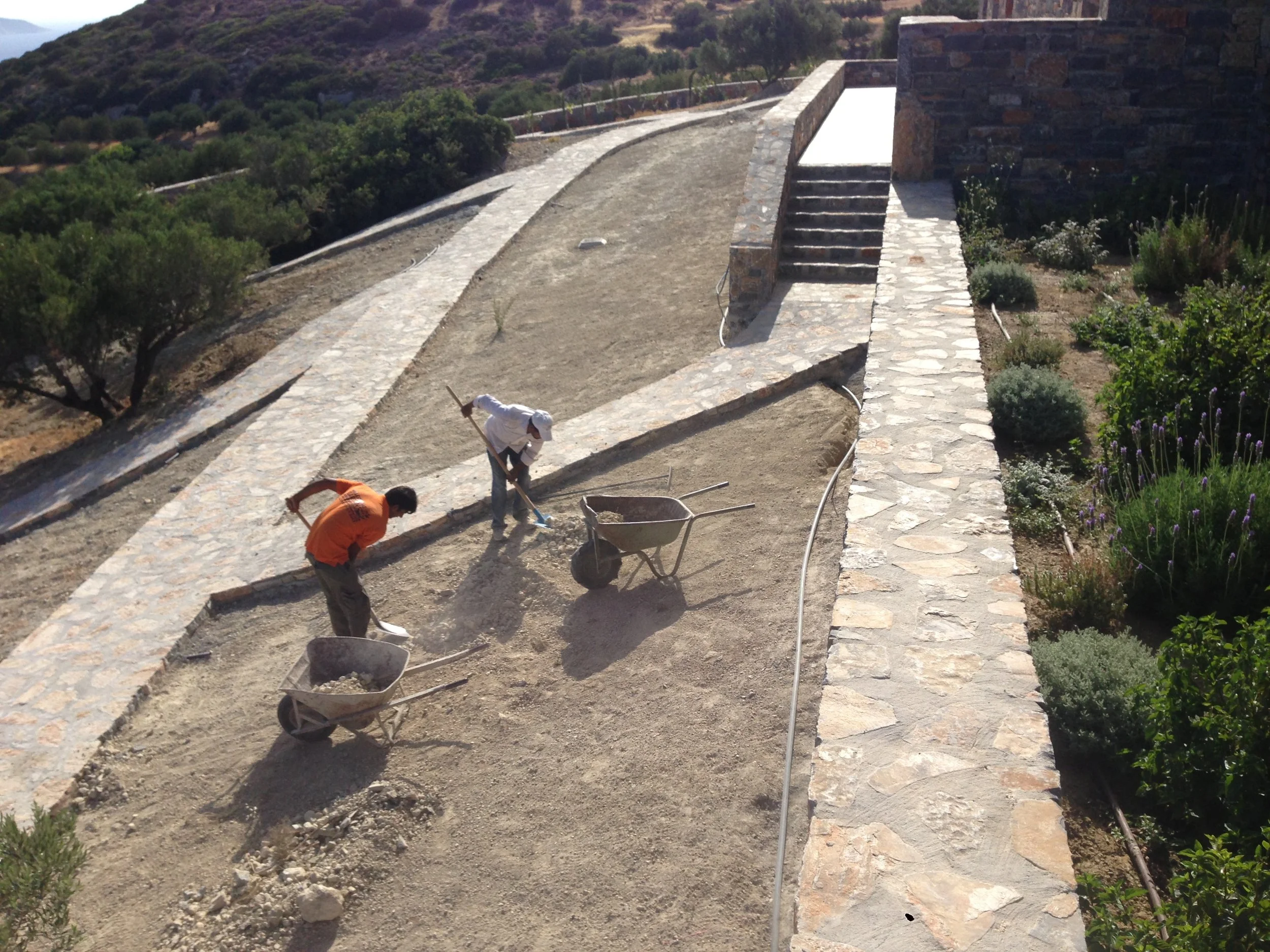 Two workers planting or working on a garden pathway with stone borders in a hilly area, with trees and plants growing nearby.