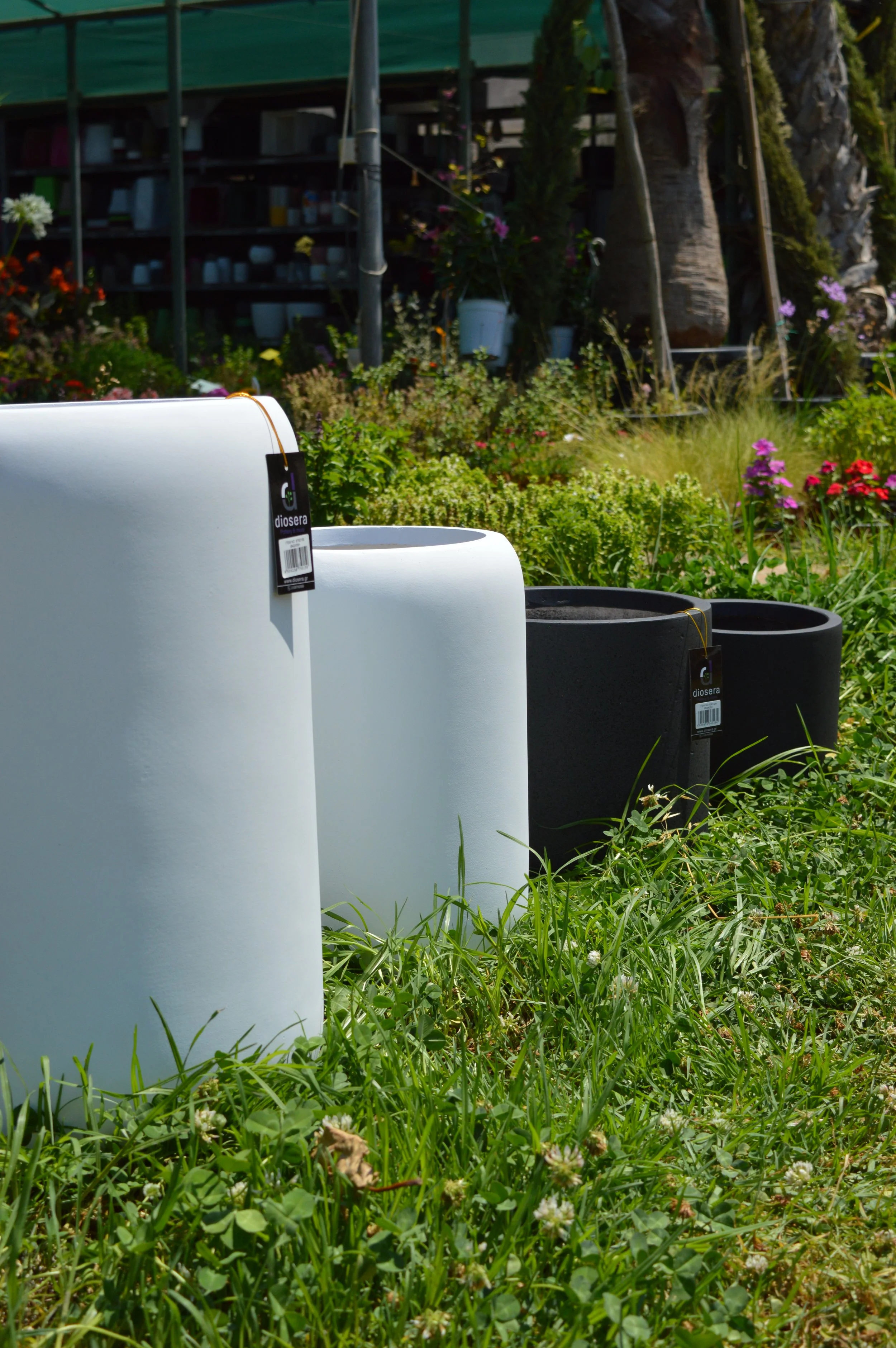 Close-up view of three large flower pots placed on grass, with a garden in the background including trees, flowers, and a structure with shelves.