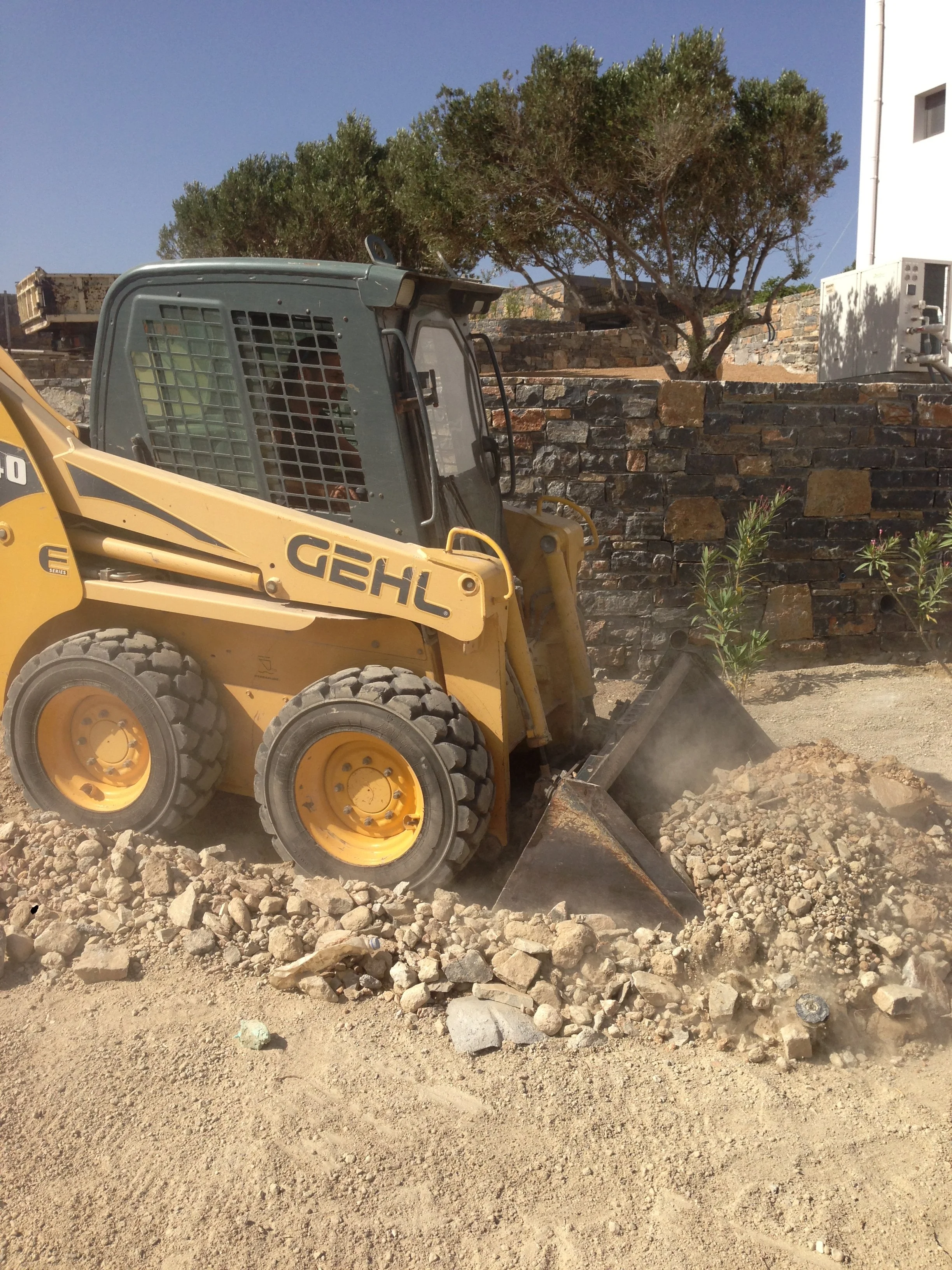 A yellow construction vehicle, a skid steer loader, is moving dirt and rocks at a construction site. In the background, there is a brick wall, a tree, and a building with an electrical unit.