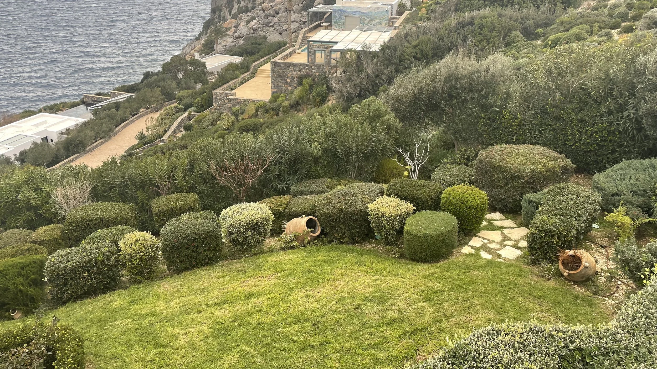 Lush garden with trimmed bushes and trees on a hillside overlooking the ocean. Stone pathway and decorative pots are visible, with modern buildings on the slope in the background.