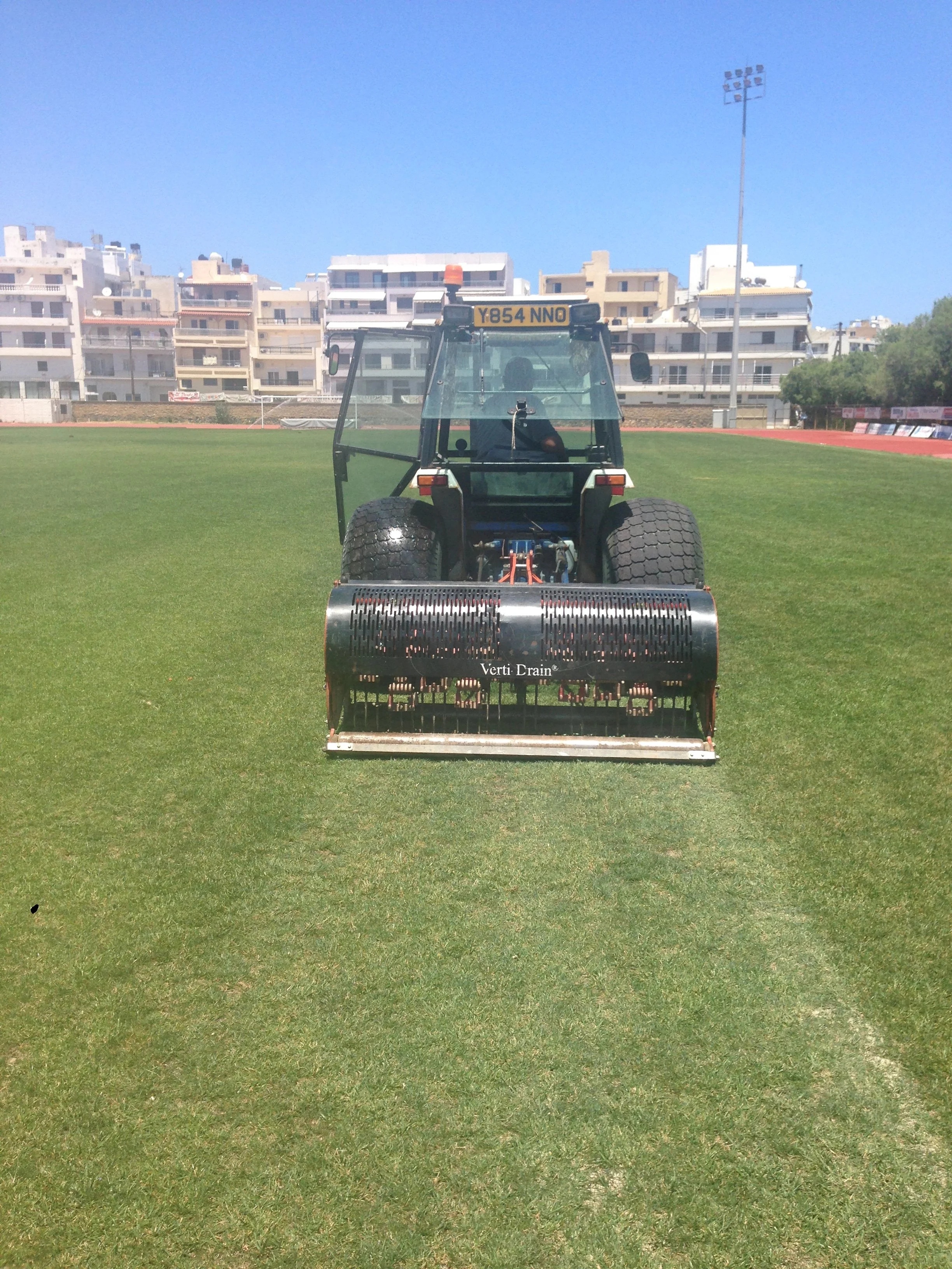 A turf maintenance machine on a sports field with a cityscape of apartment buildings and a light pole in the background.