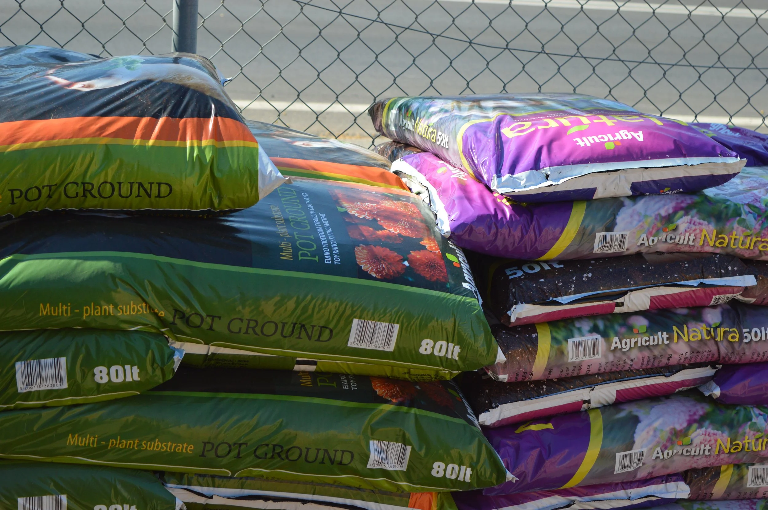 Stacked bags of potting soil labeled 'Pot Ground' and 'Agricul Natural' in front of a chain-link fence.