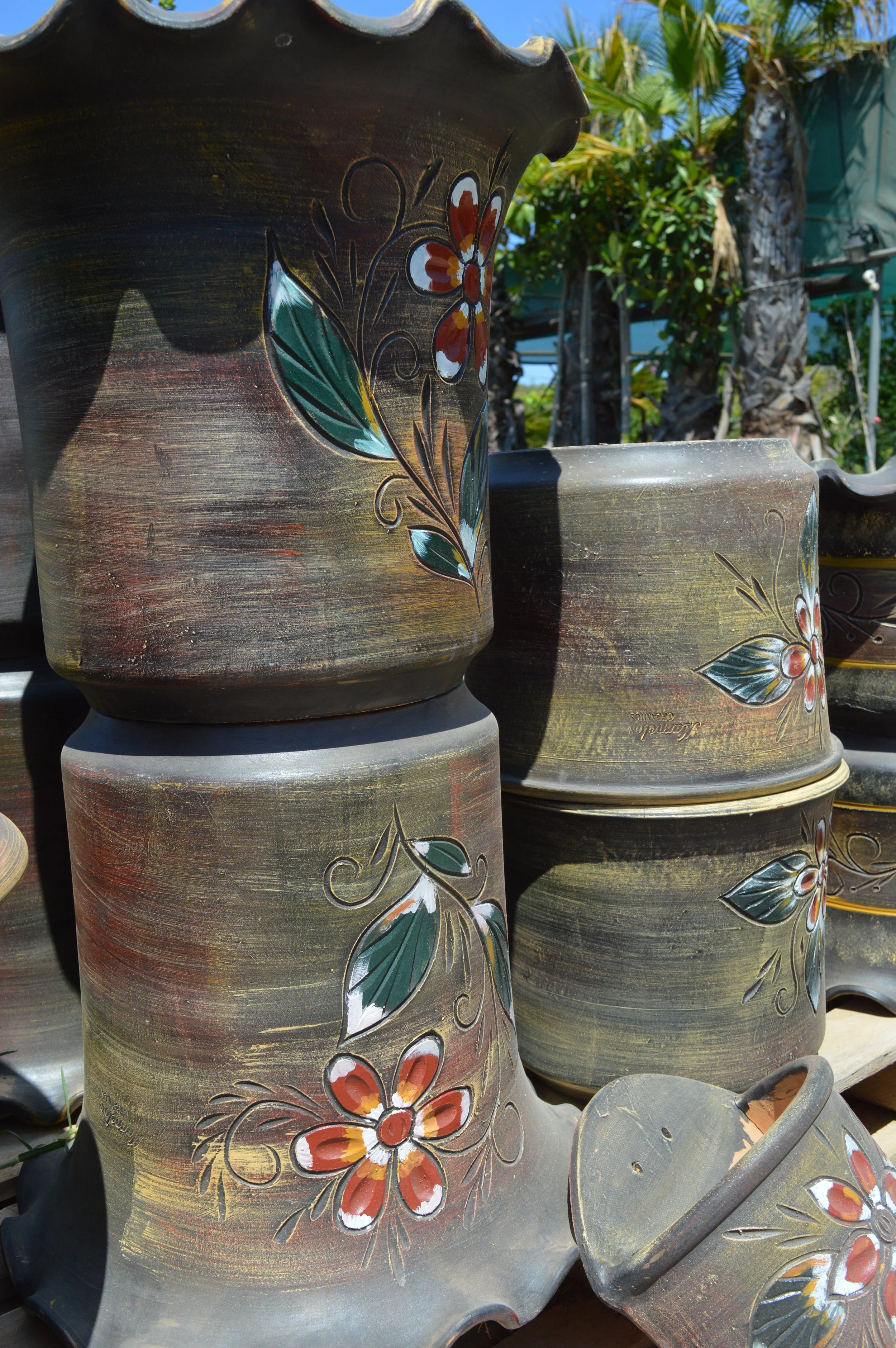 Multiple rustic brown flower pots decorated with colorful painted flowers, outdoors with green trees and blue sky in the background.