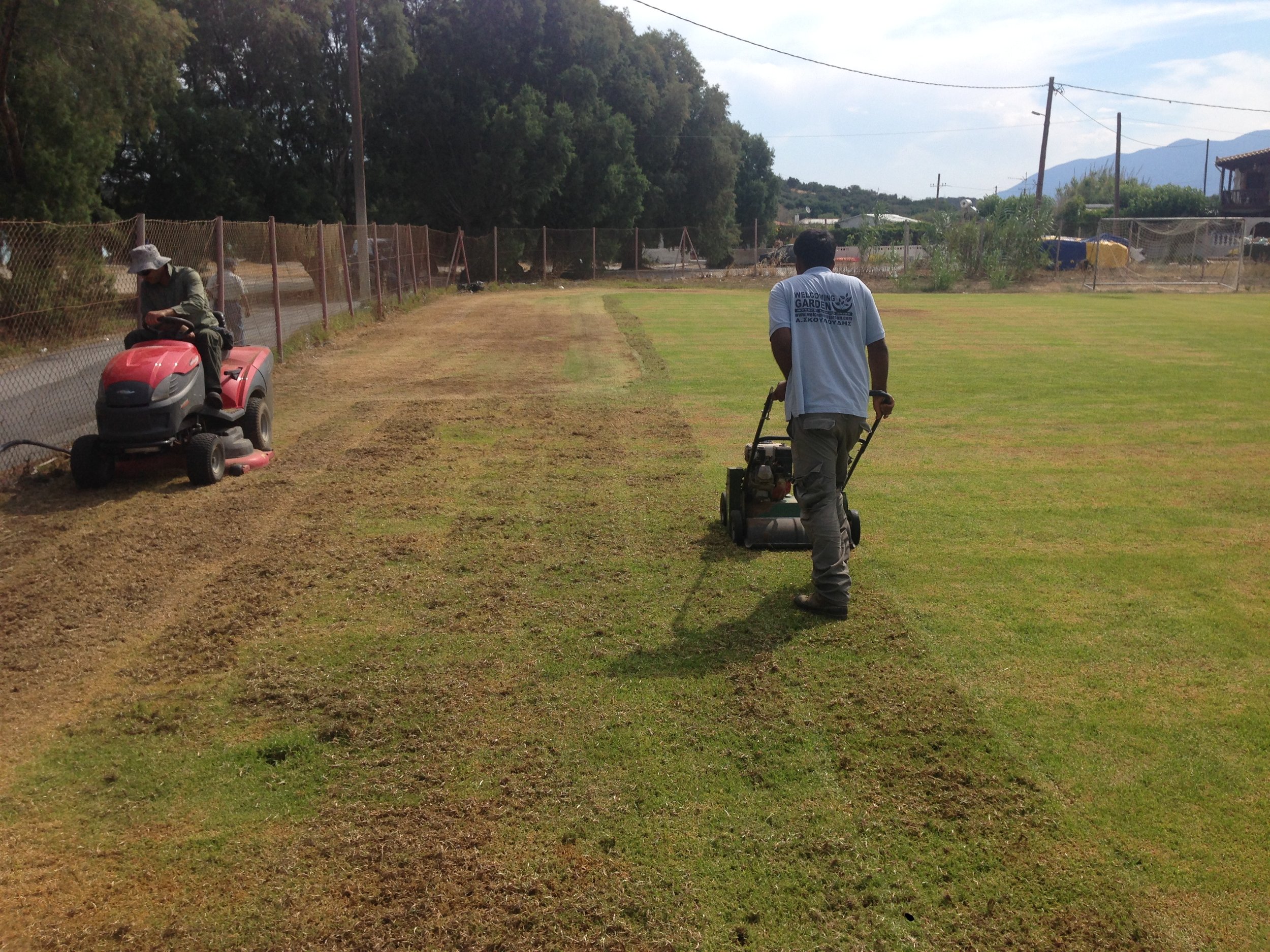 People working on a lawn, with one person riding a red riding lawn mower and another using a push mower, bordered by a fence with trees and mountains in the background.