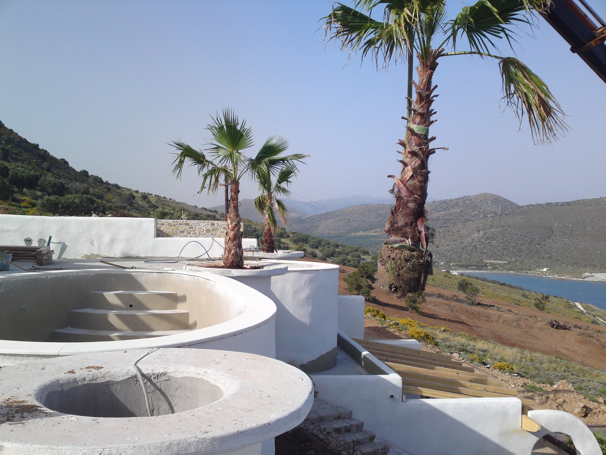 Construction site with white buildings, palm trees, and a scenic hilly landscape with a body of water in the background.