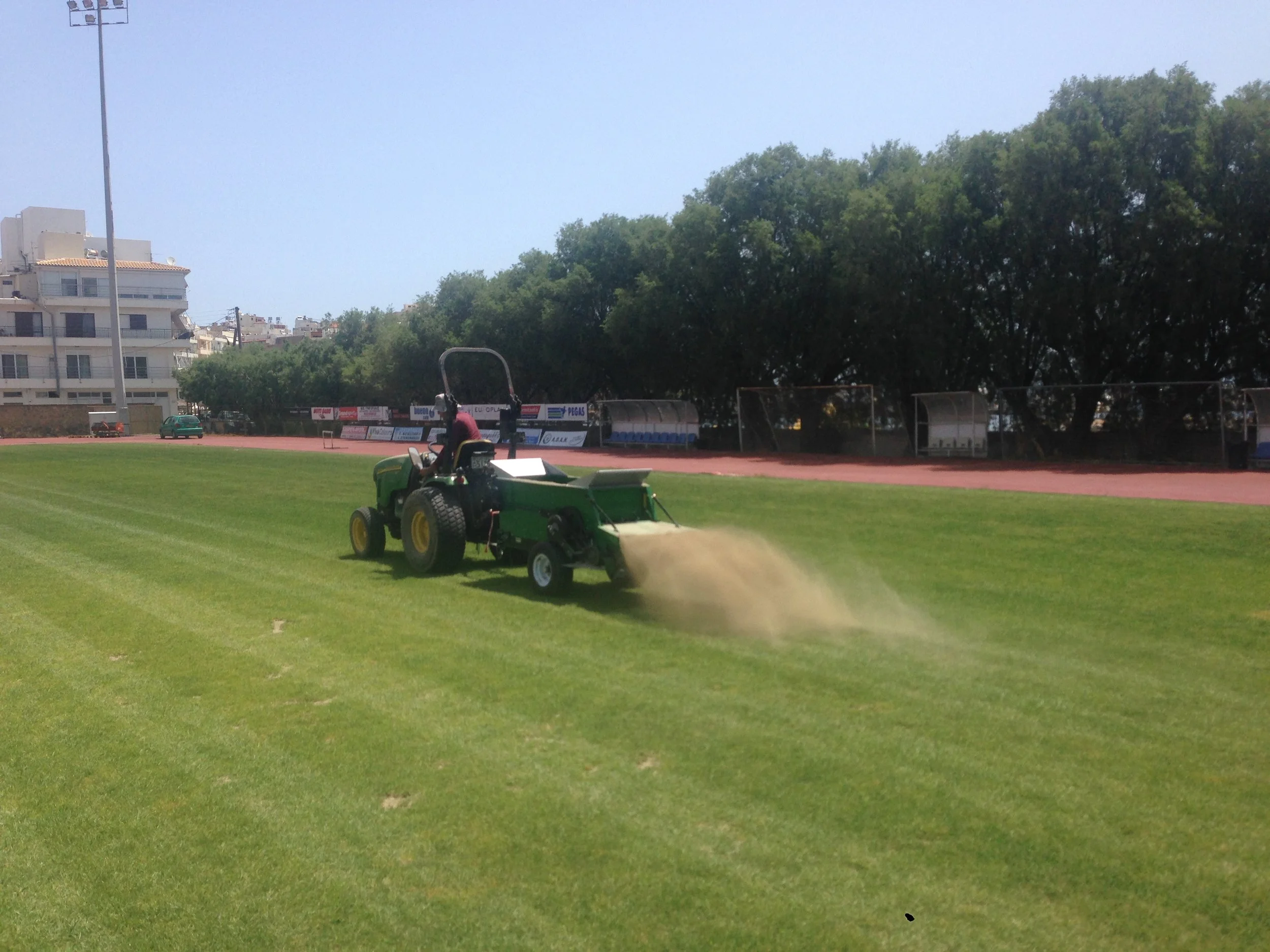 A green riding lawn mower on a sports field, watering the grass with a sprinkler attachment on a sunny day.