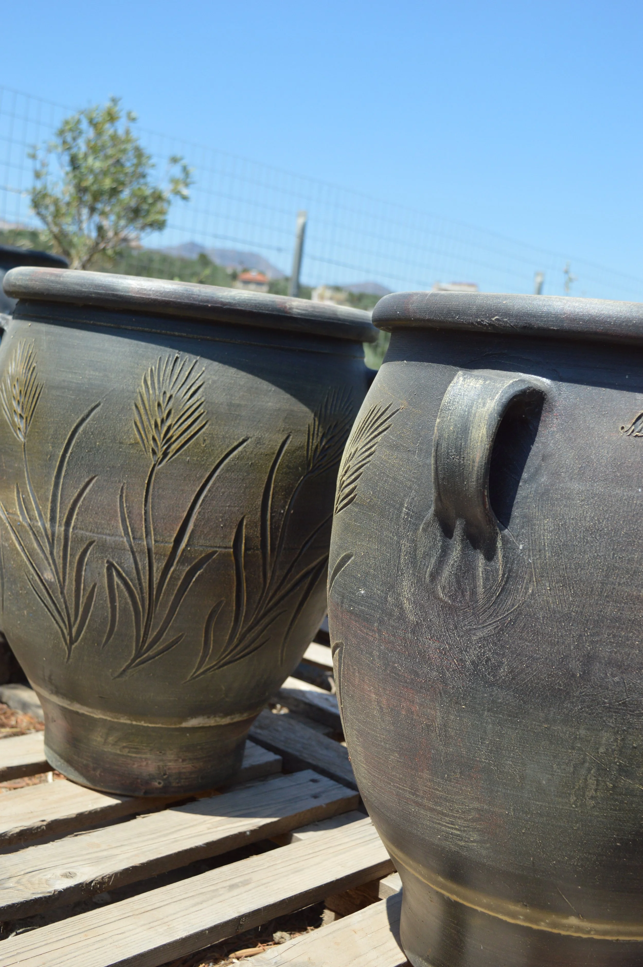 Close-up of three decorative outdoor plant pots with engraved leaf patterns, placed on a wooden pallet against a backdrop of a wire fence, trees, and blue sky.