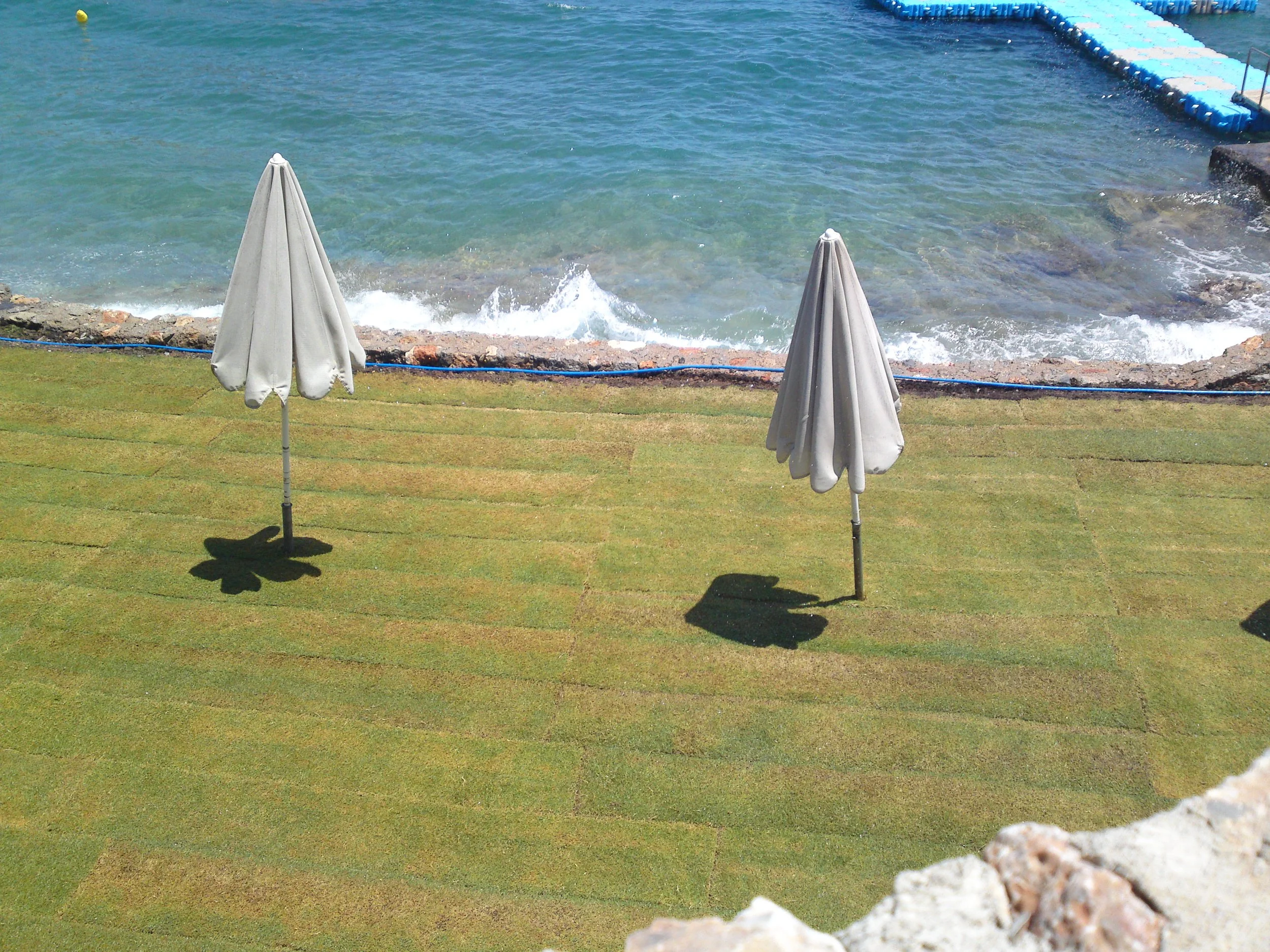 A view of the ocean with waves crashing against a rocky shoreline. Two closed white umbrellas are on a grassy area in the foreground. There is a floating barrier in the water, and the sky appears clear.