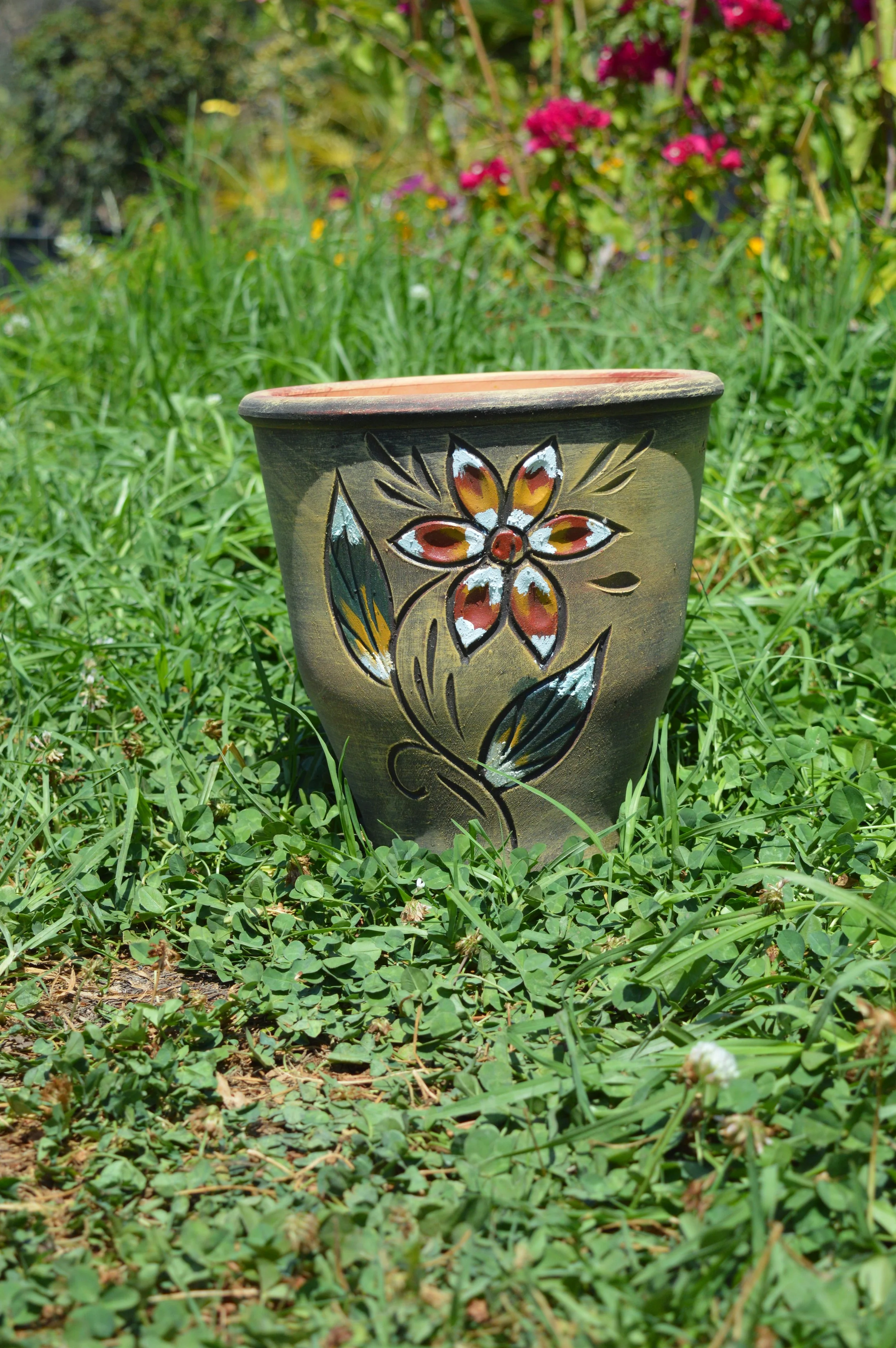 A painted ceramic flowerpot with colorful floral designs sitting on grass in a garden with various green plants and pink flowers in the background.