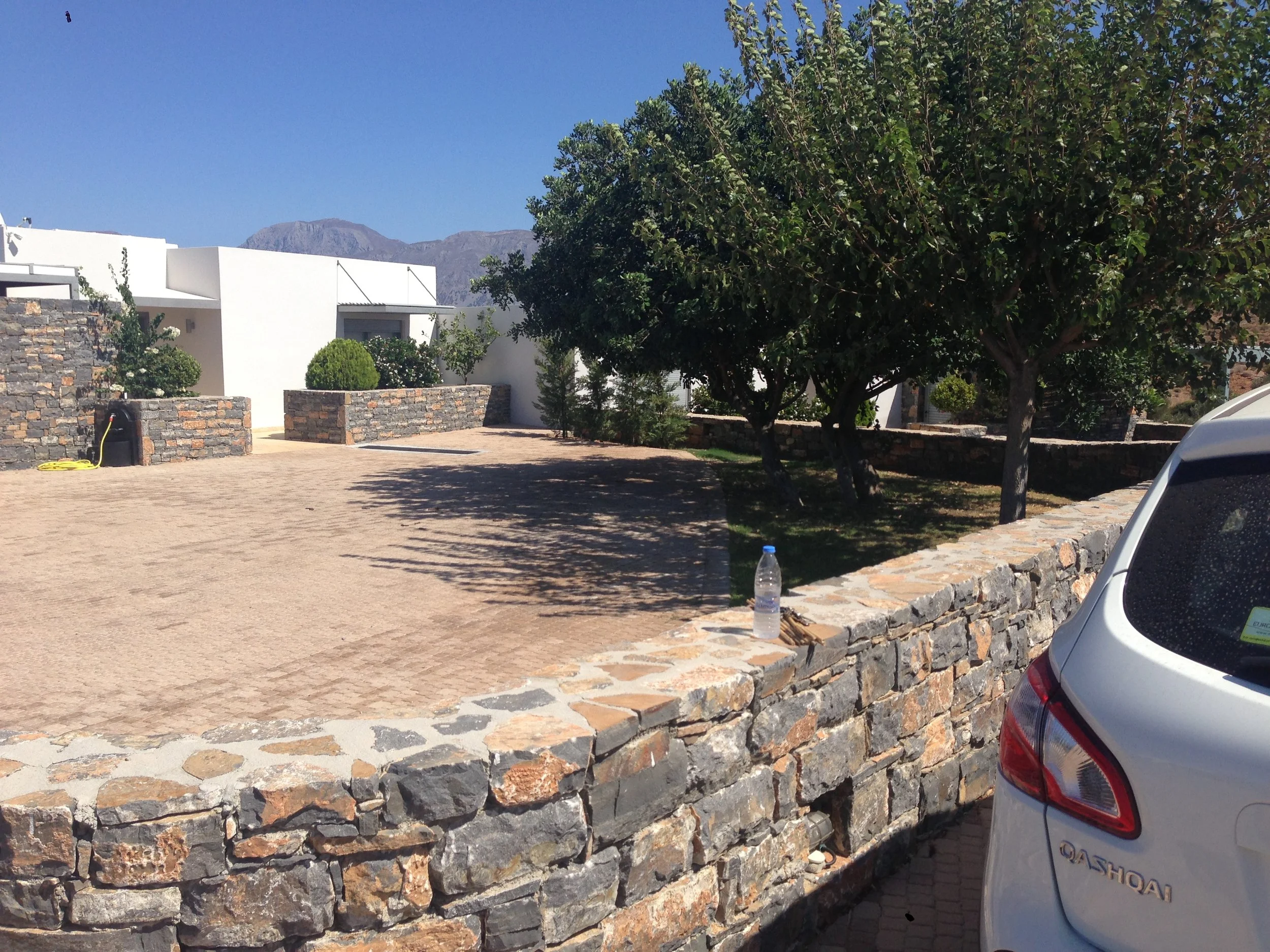A residential area with white modern houses, stone walls, a big tree, and mountains in the background. Part of a silver car is visible on the right.