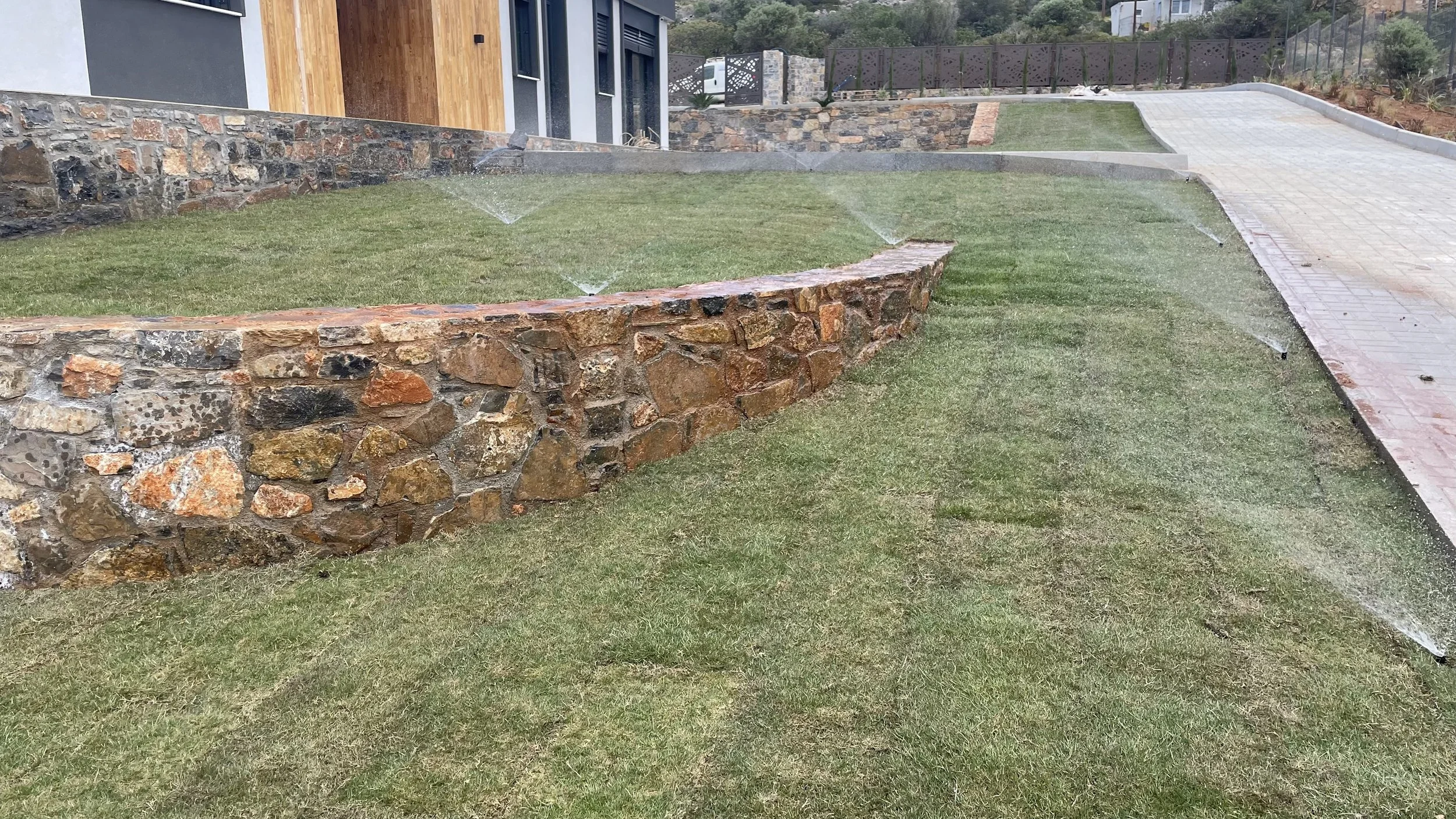 A landscaped yard featuring a stone retaining wall with water sprinklers, a grassy area, a paved walkway, and a modern house in the background.