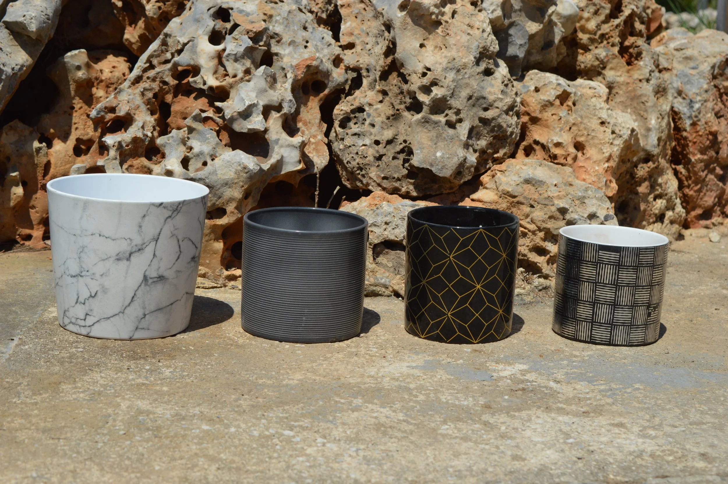 Four decorative mugs with various black and white patterns placed on the ground in front of large, porous rocks.