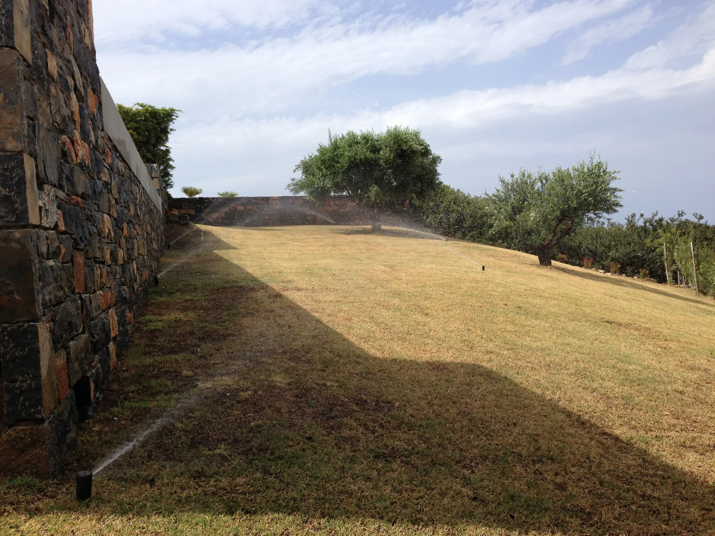 A landscaped yard with a stone wall on the left side, green grass, and trees in the background, under a partially cloudy sky.