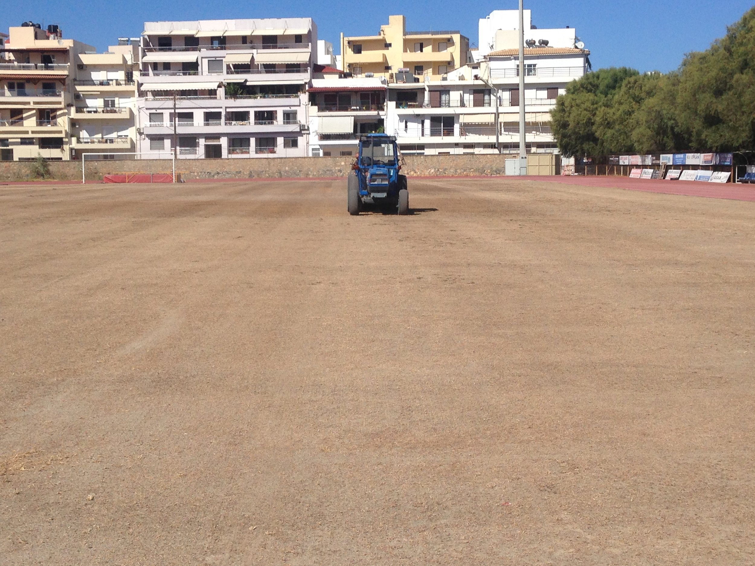 A blue tractor on a dirt field in front of residential buildings and trees under a clear blue sky.