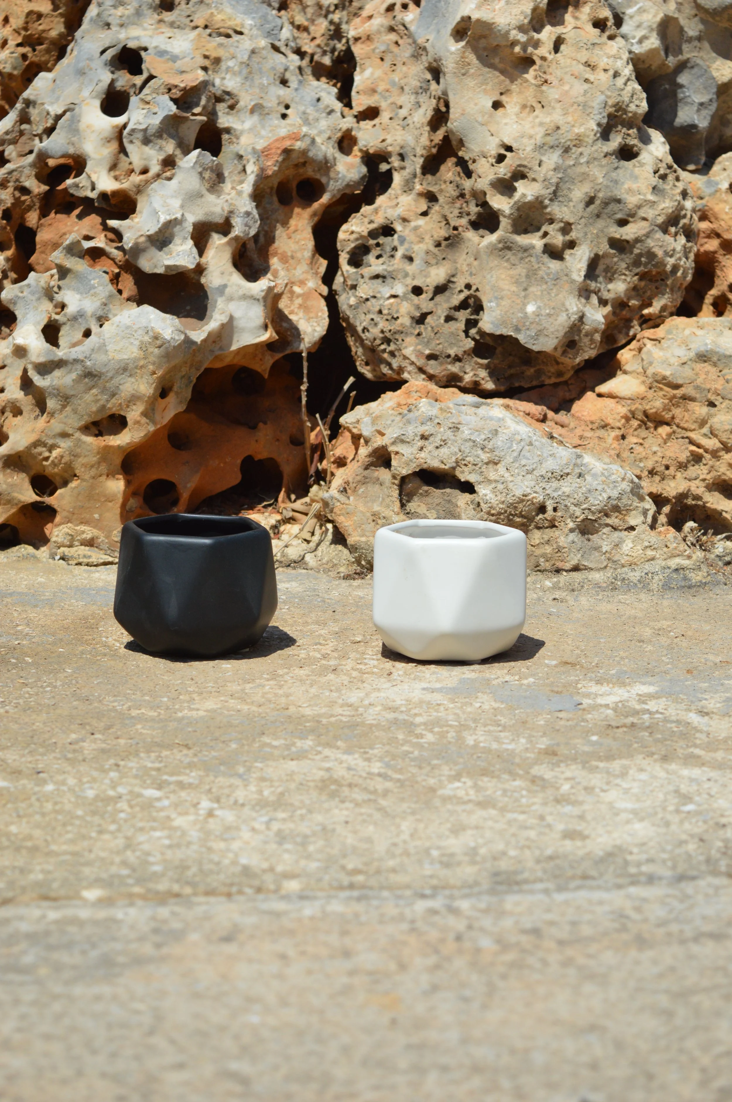 Close-up of a black and white ceramic mug on a rough sandy surface, with a rocky background featuring textured rocks with holes.