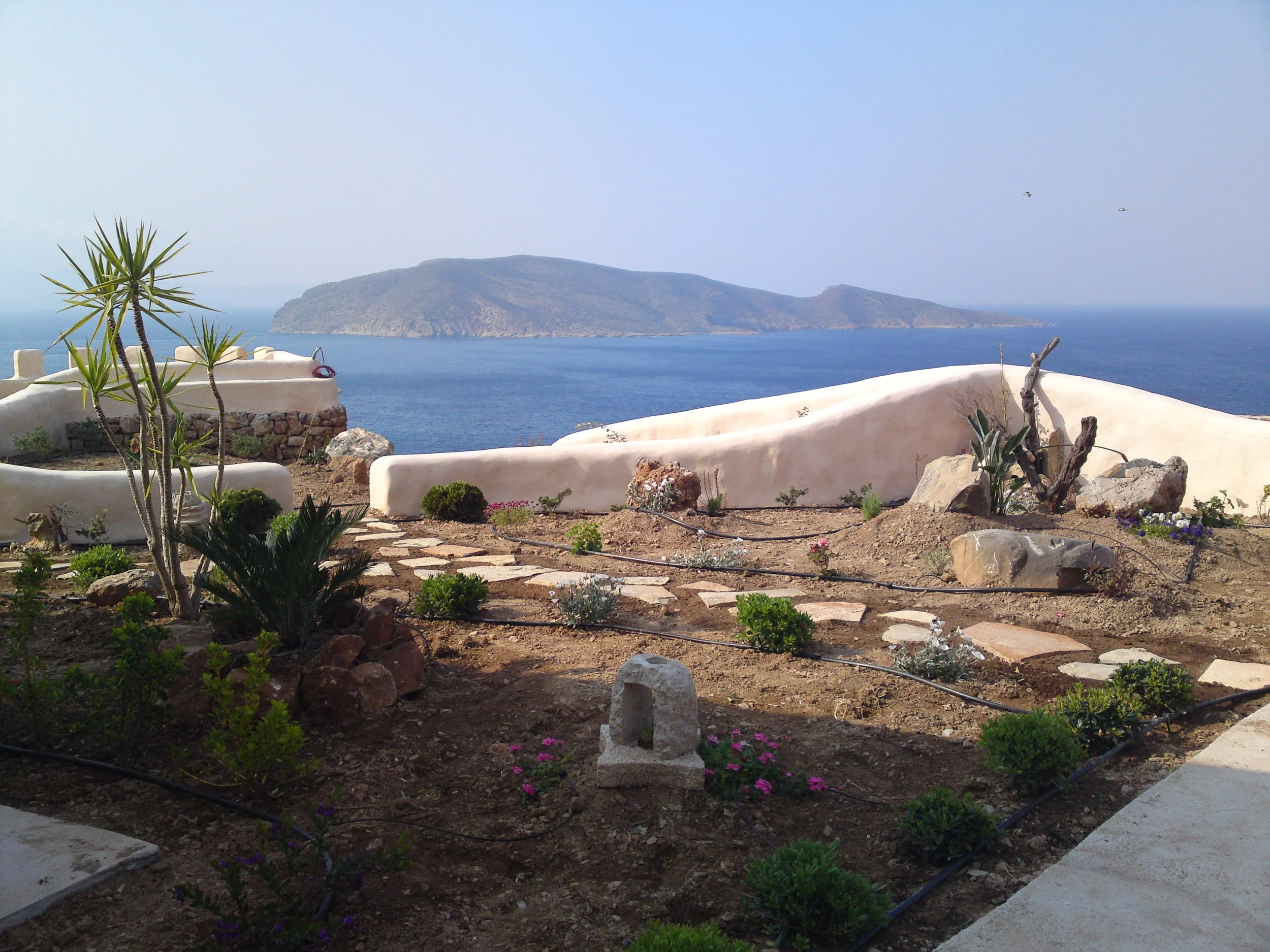 A seaside garden with small plants and flowers in the foreground, old stone structures and a dirt path, a white curved wall, and an ocean with an island and two birds flying in the background.