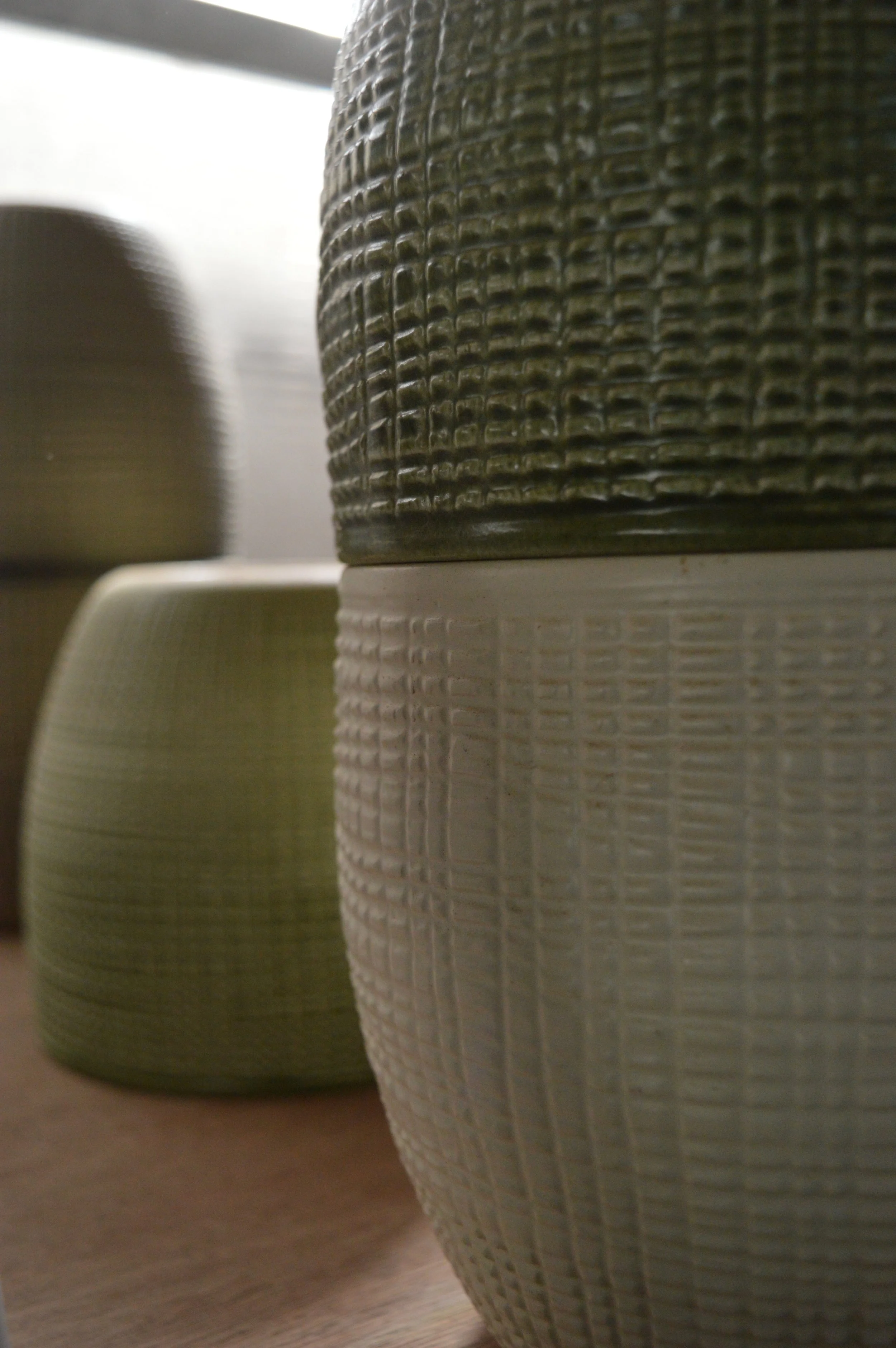 Close-up of two ceramic vases with textured patterns, one in a darker earthy tone and the other in a lighter, off-white color, placed on a wooden surface.