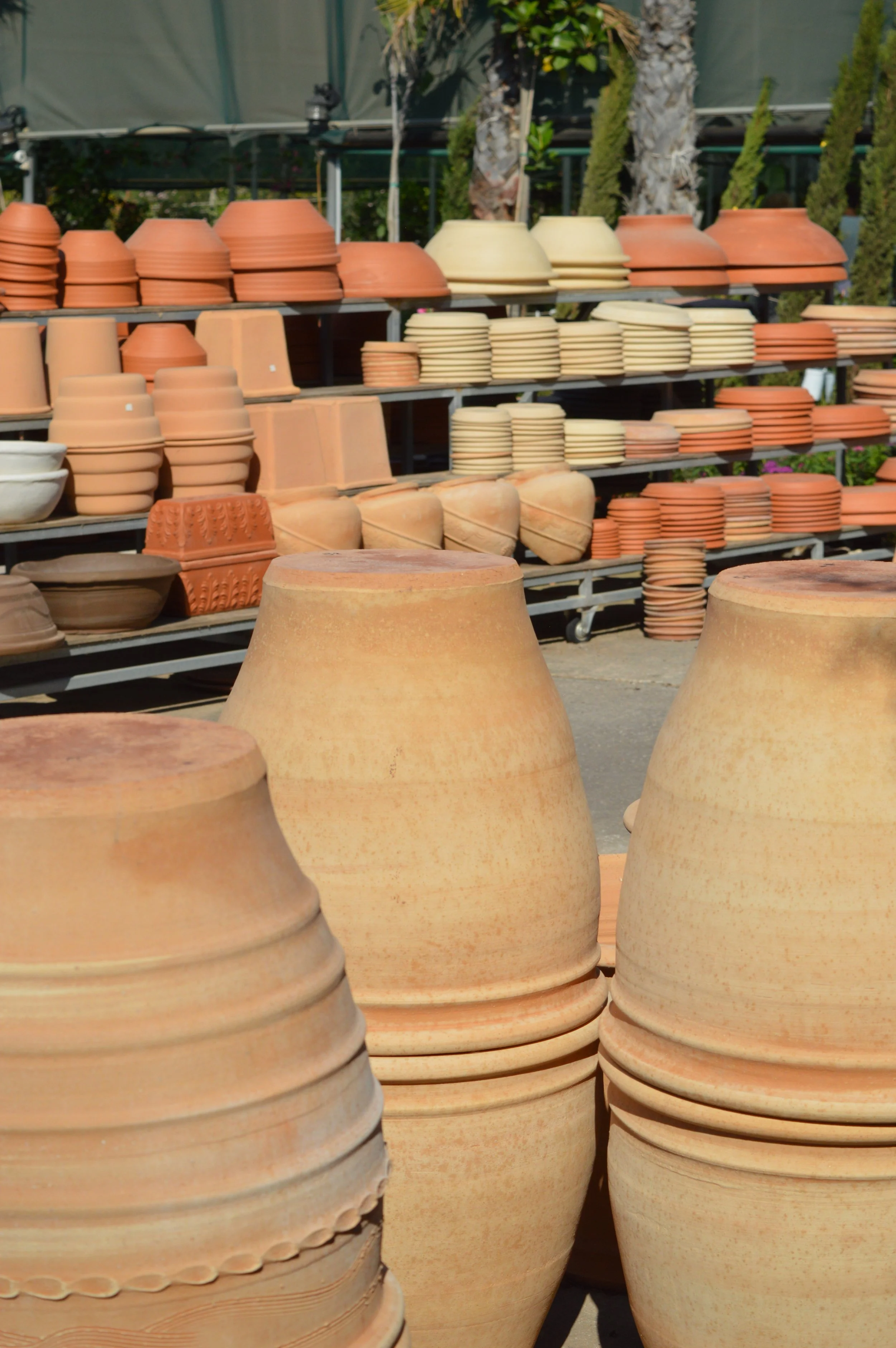 Multiple stacked terracotta and beige clay plant pots on metal shelves outdoors, with trees and greenery in the background.