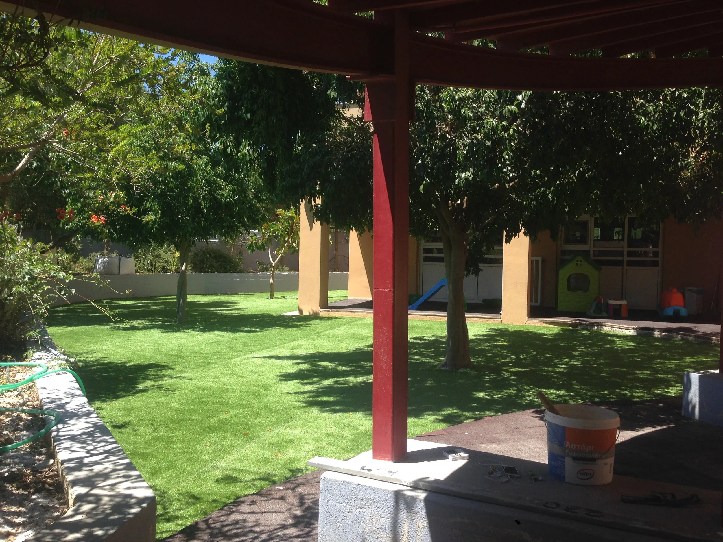 A backyard with green grass, trees, and a yellow house in the background. There is a red vertical beam supporting a wooden pergola. Various children's toys and gardening equipment are visible, including a bucket, a slide, and a small playhouse.